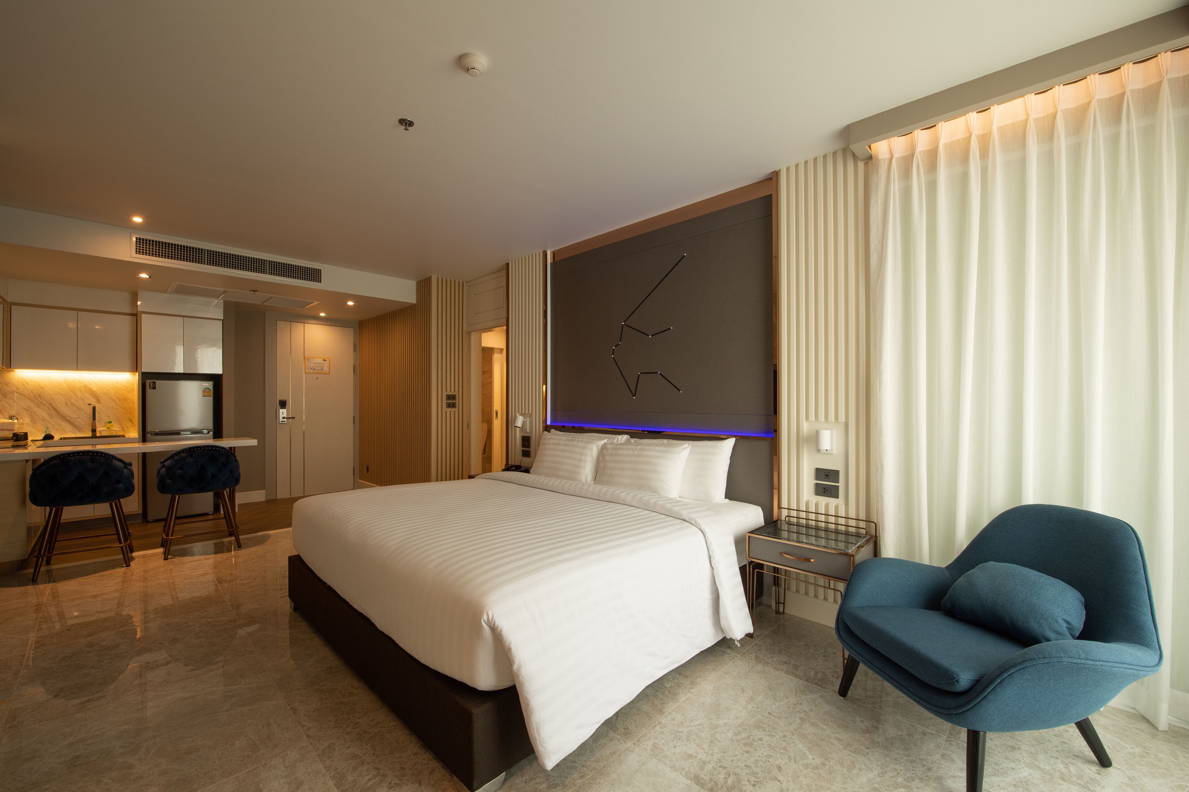 A modern hotel room with a large bed, a desk with two chairs, a small table, and a blue armchair.