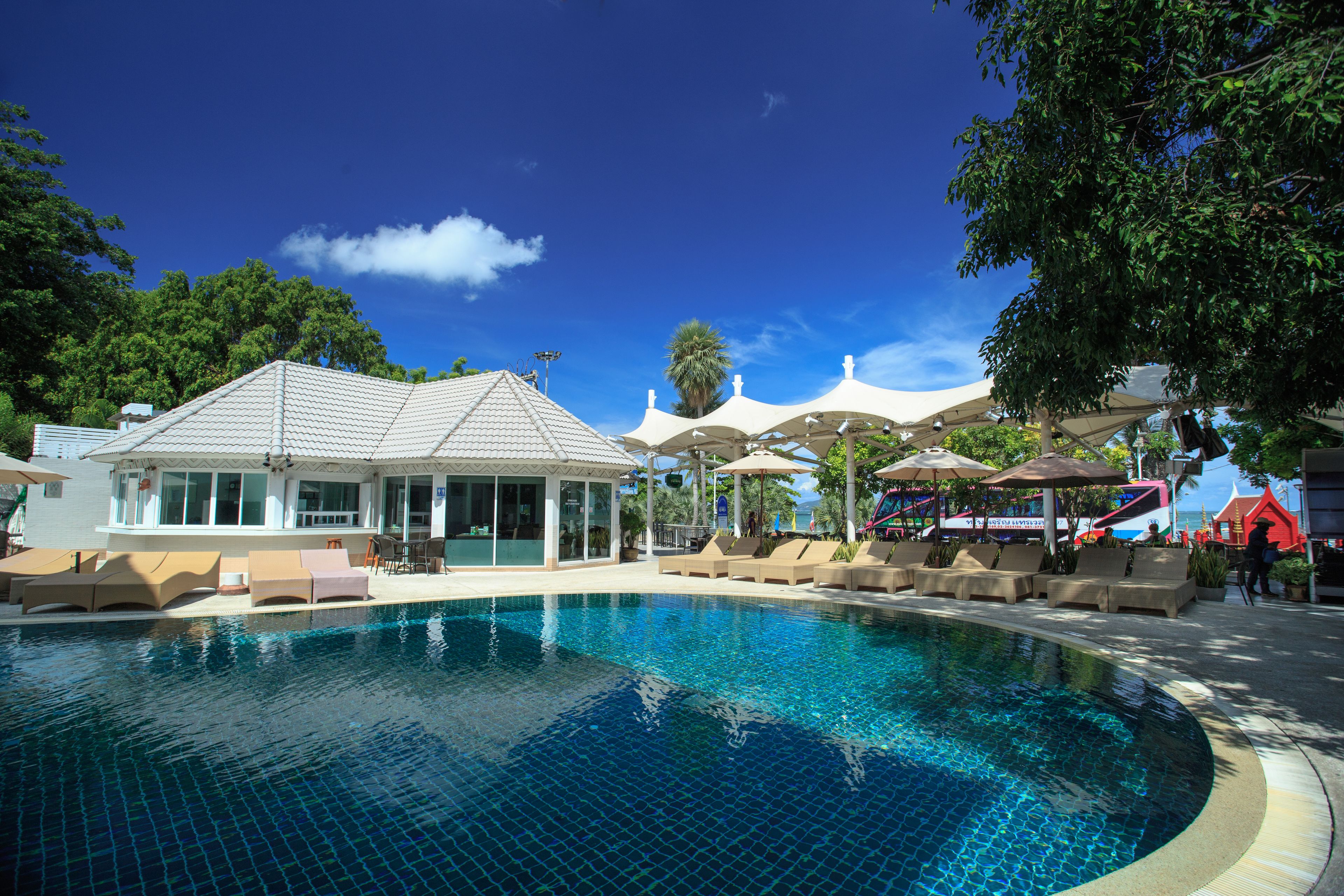Piscina com espreguiçadeiras e guarda-sóis, rodeada por um edifício com telhado de azulejos e área de estar coberta.