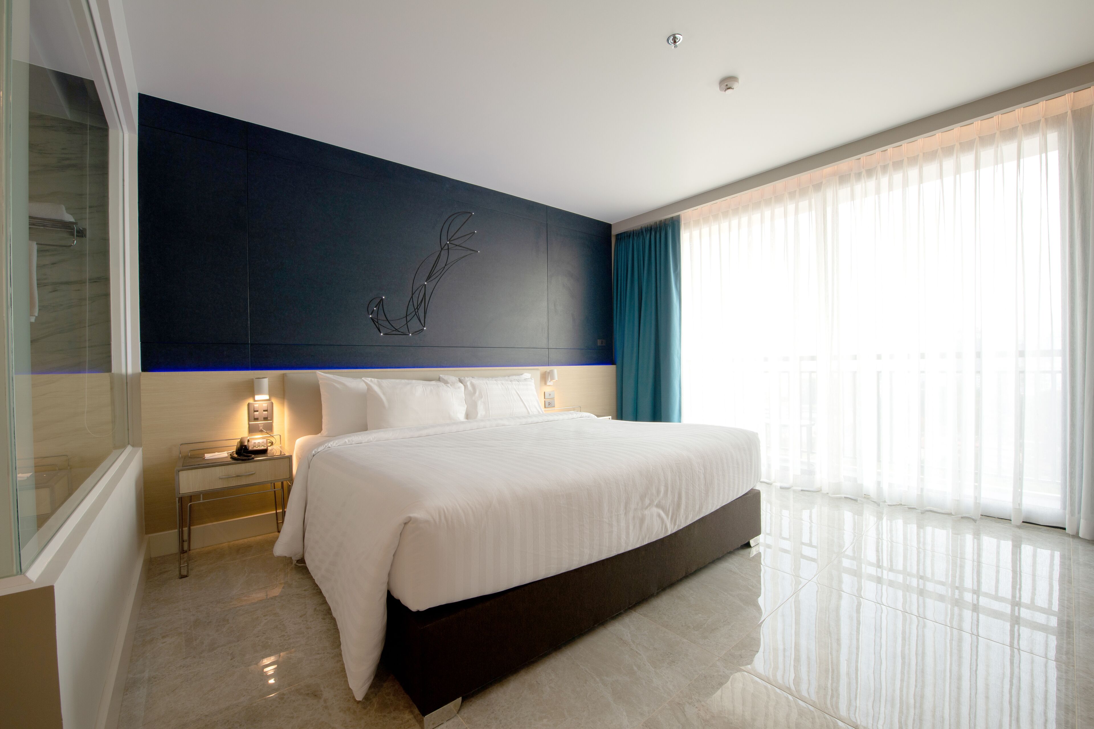 A modern hotel room with a large bed, a bedside table with a lamp, a wall-mounted artwork, and a large window with curtains.