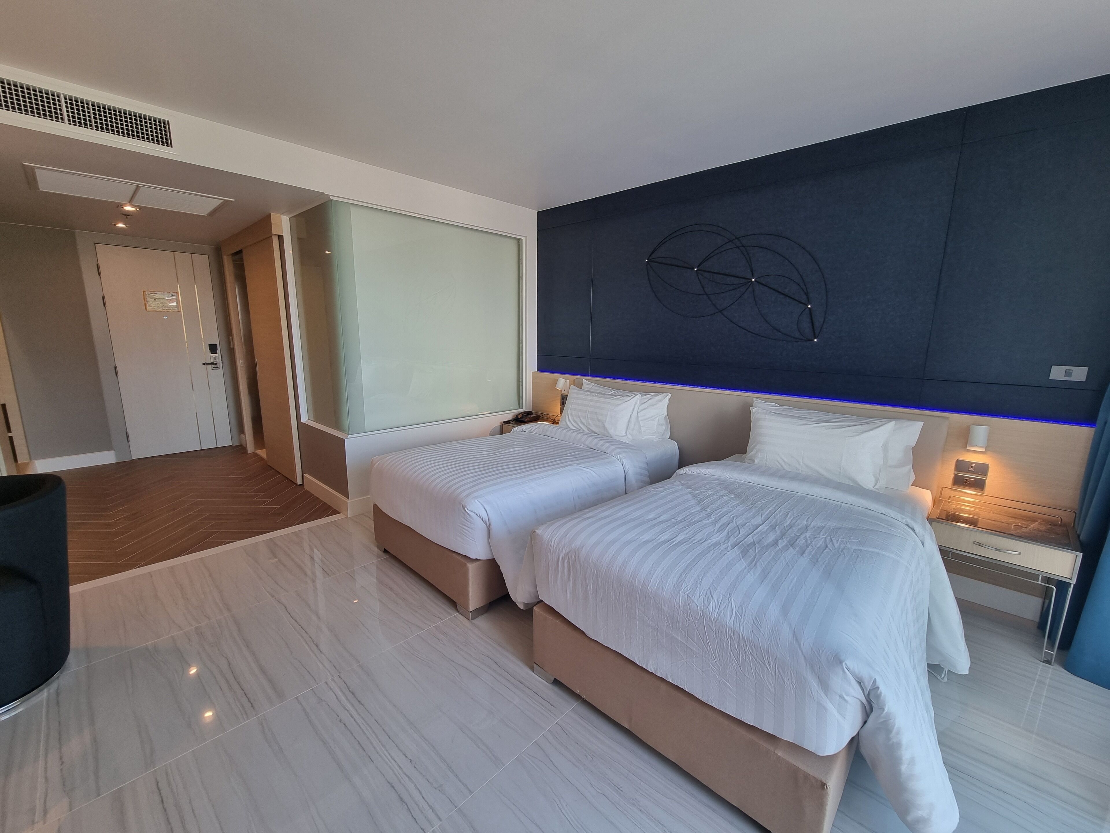 Une chambre d’hôtel avec deux lits, une grande fenêtre, une tête de lit moderne et une table de chevet avec une lampe.