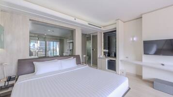 A modern hotel room with a large bed, a television, a minibar, and a bathroom with a glass shower enclosure.