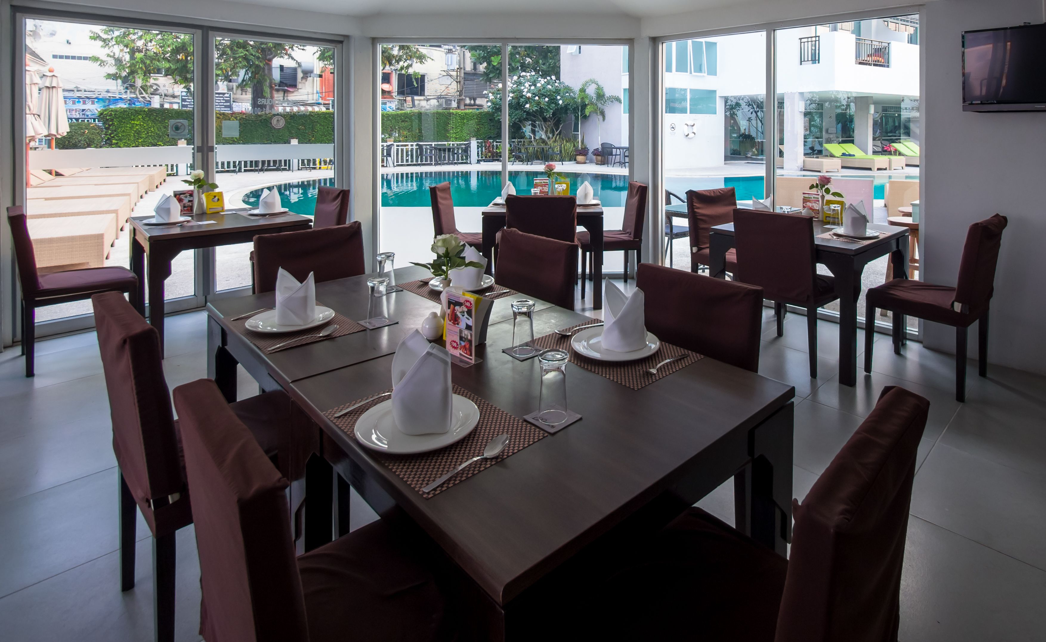 A dining area with tables set for four, large windows offering views of a swimming pool and outdoor seating.