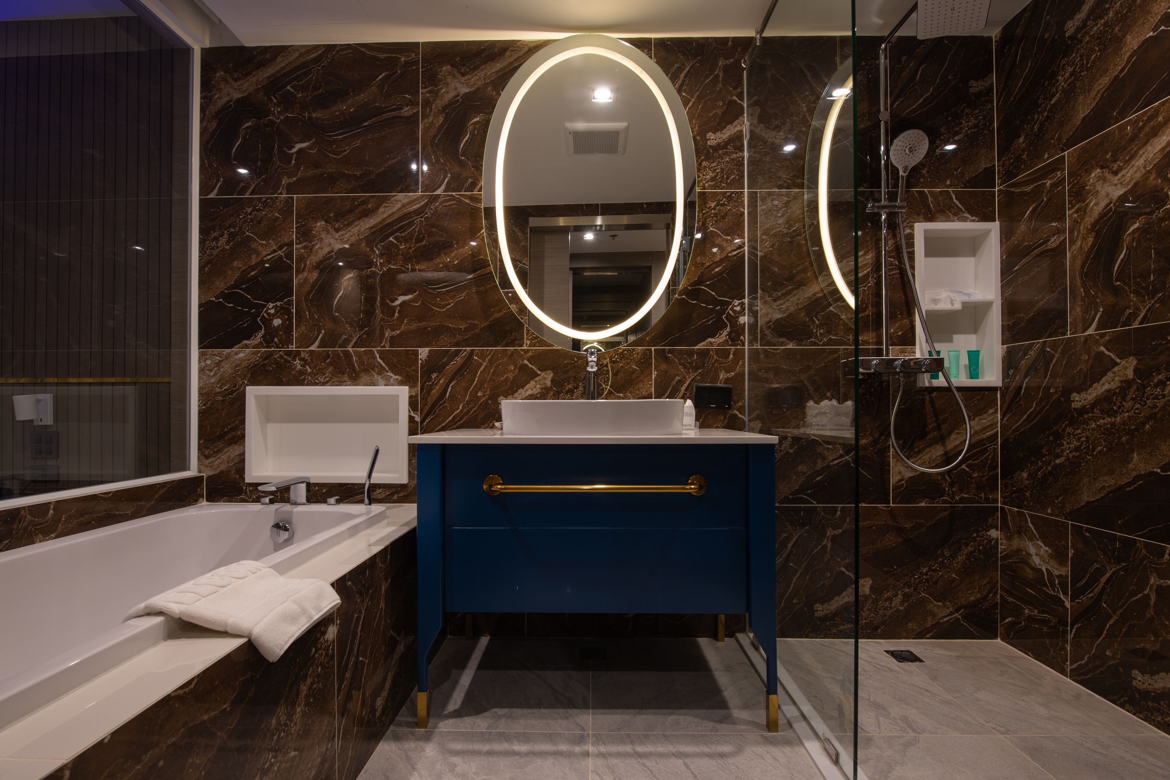 A modern bathroom with a marble wall, a white sink, a blue vanity, a glass-enclosed shower, and a large mirror with a lighted frame.