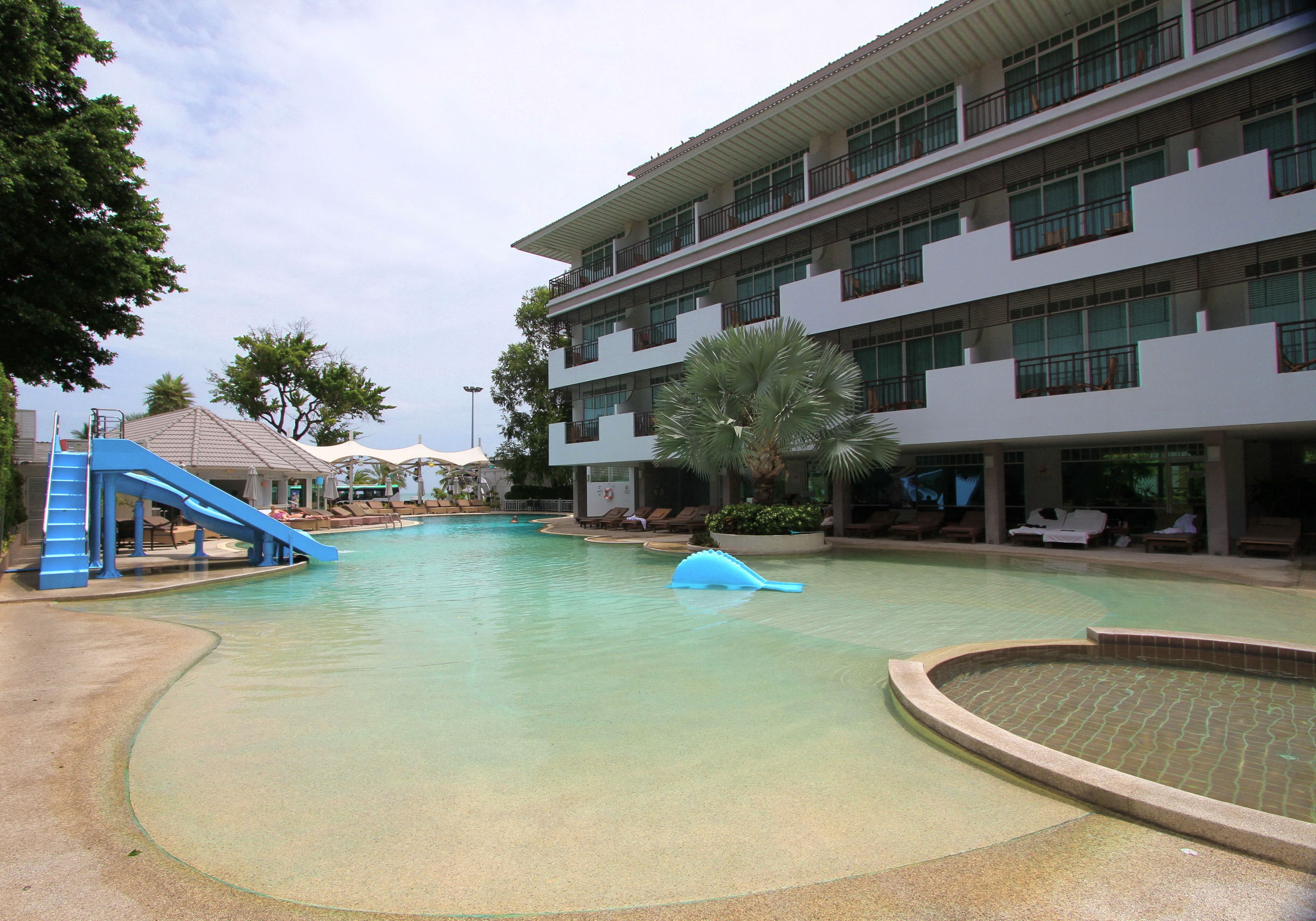 Un área de piscina con un tobogán azul, sillas de descanso y un edificio de varios pisos con balcones.