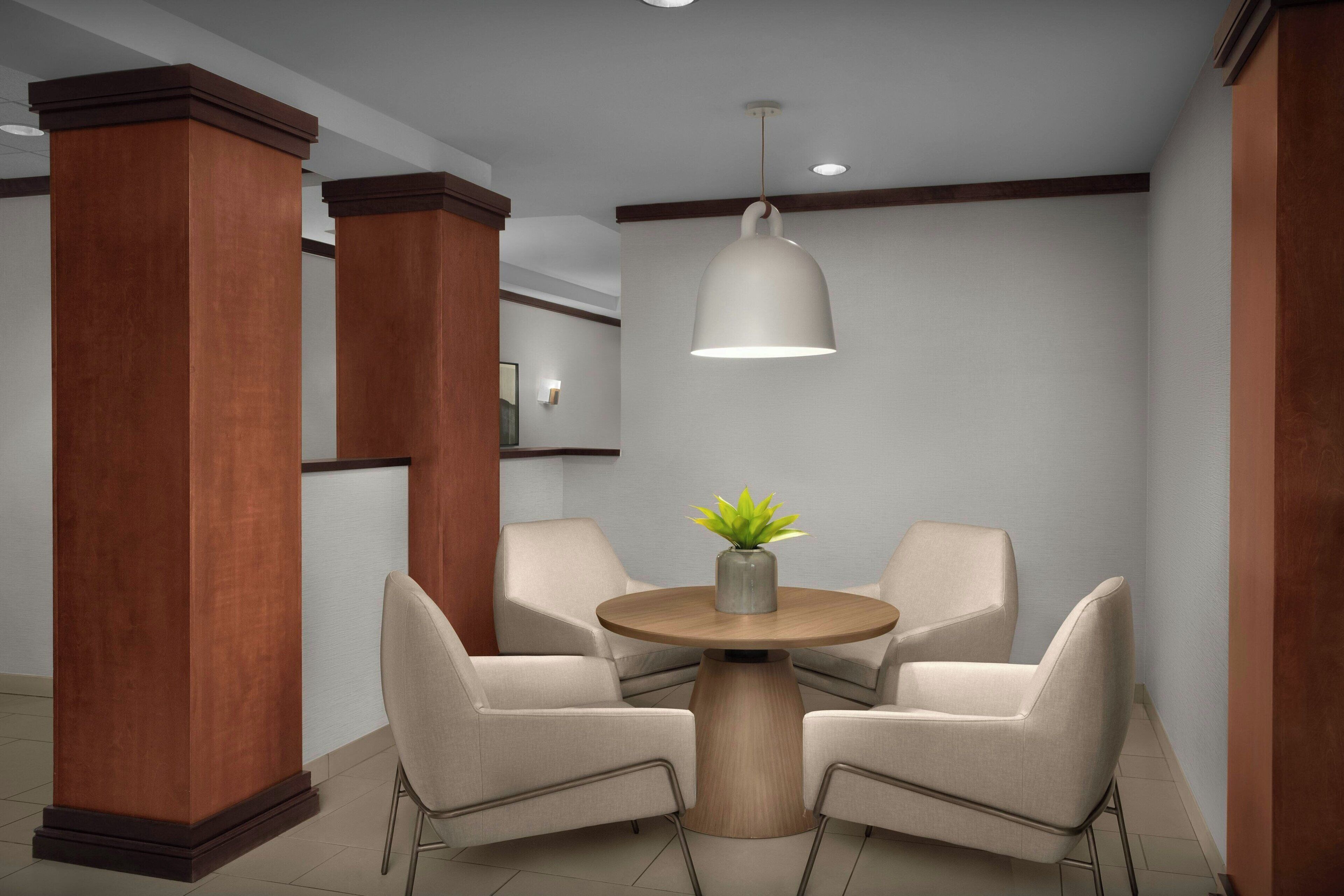 A modern waiting area with two beige chairs around a wooden table, a potted plant, and a pendant light.