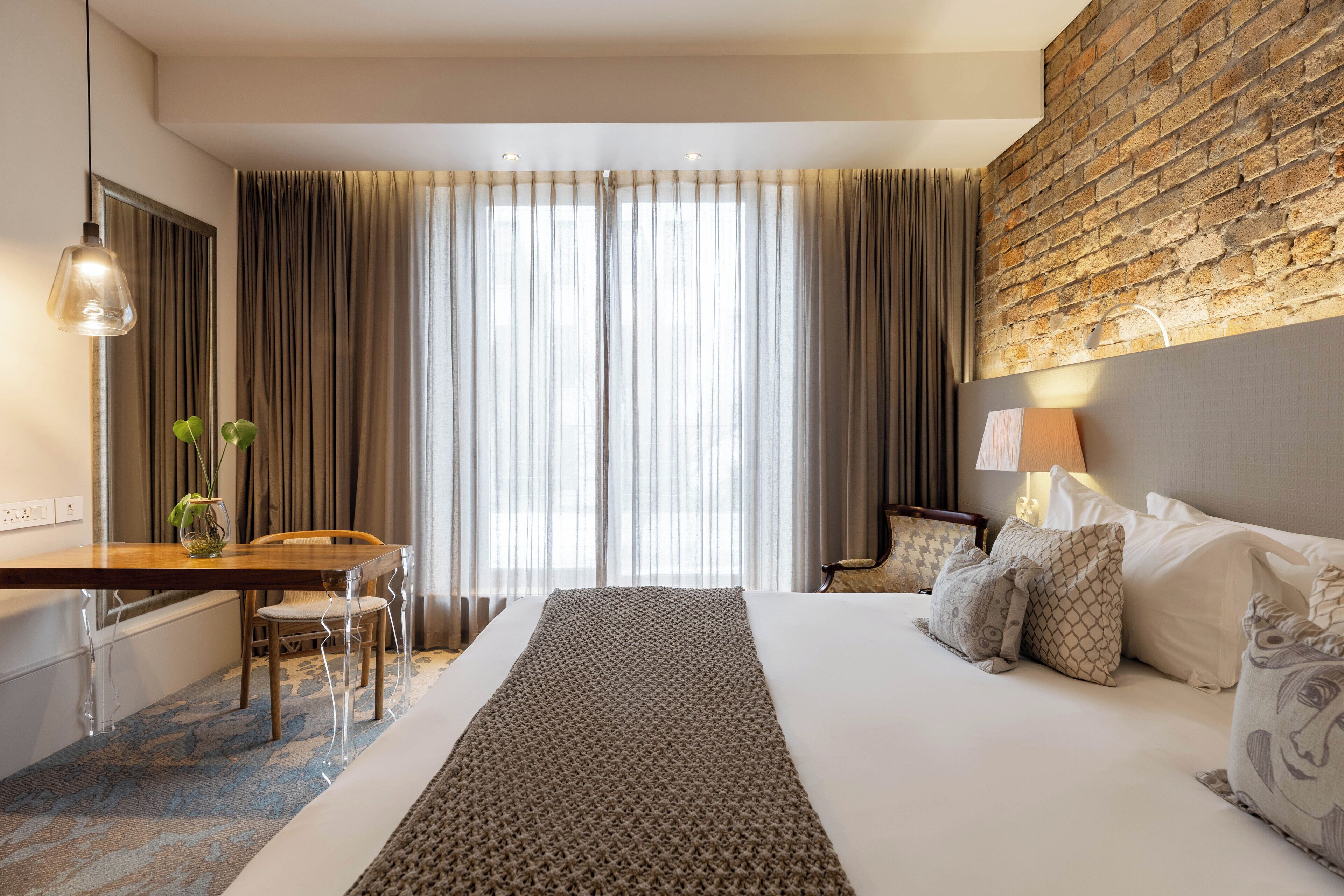 A hotel room with a large bed, a desk with a chair, a lamp, and a view of the outdoors through sheer curtains.