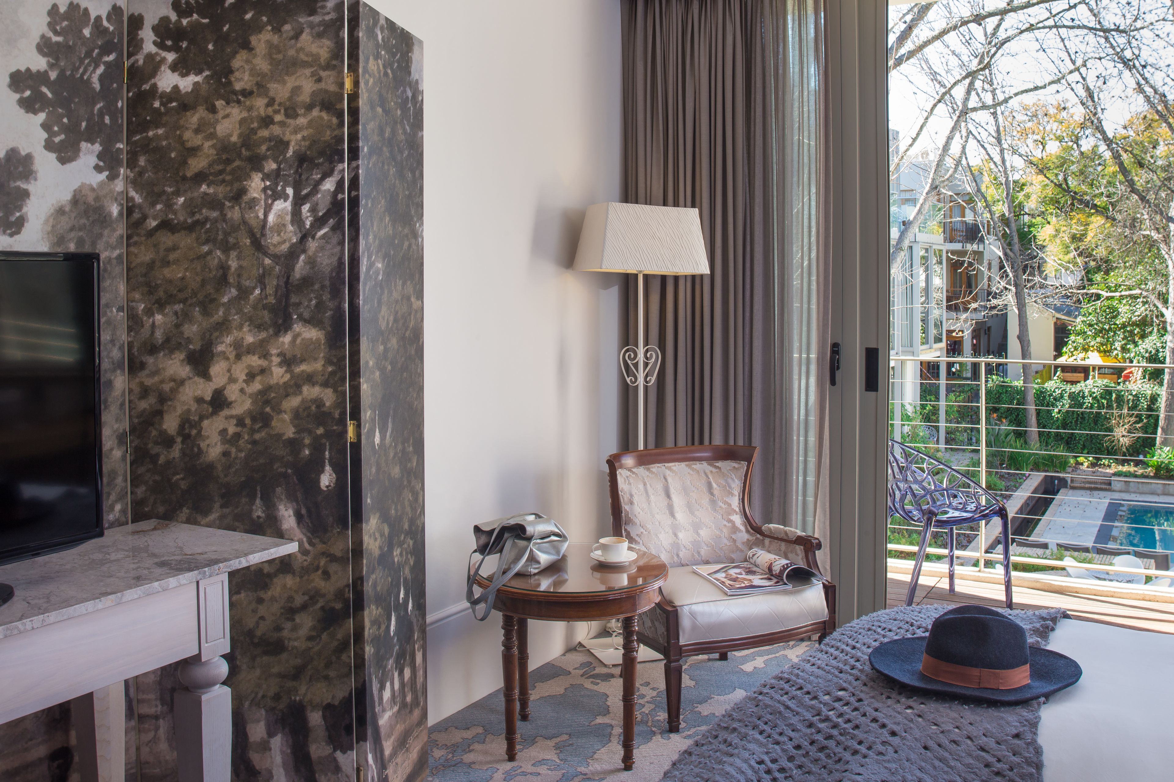A hotel room with a TV, a chair, a table with a lamp, and a view of a pool and trees outside.