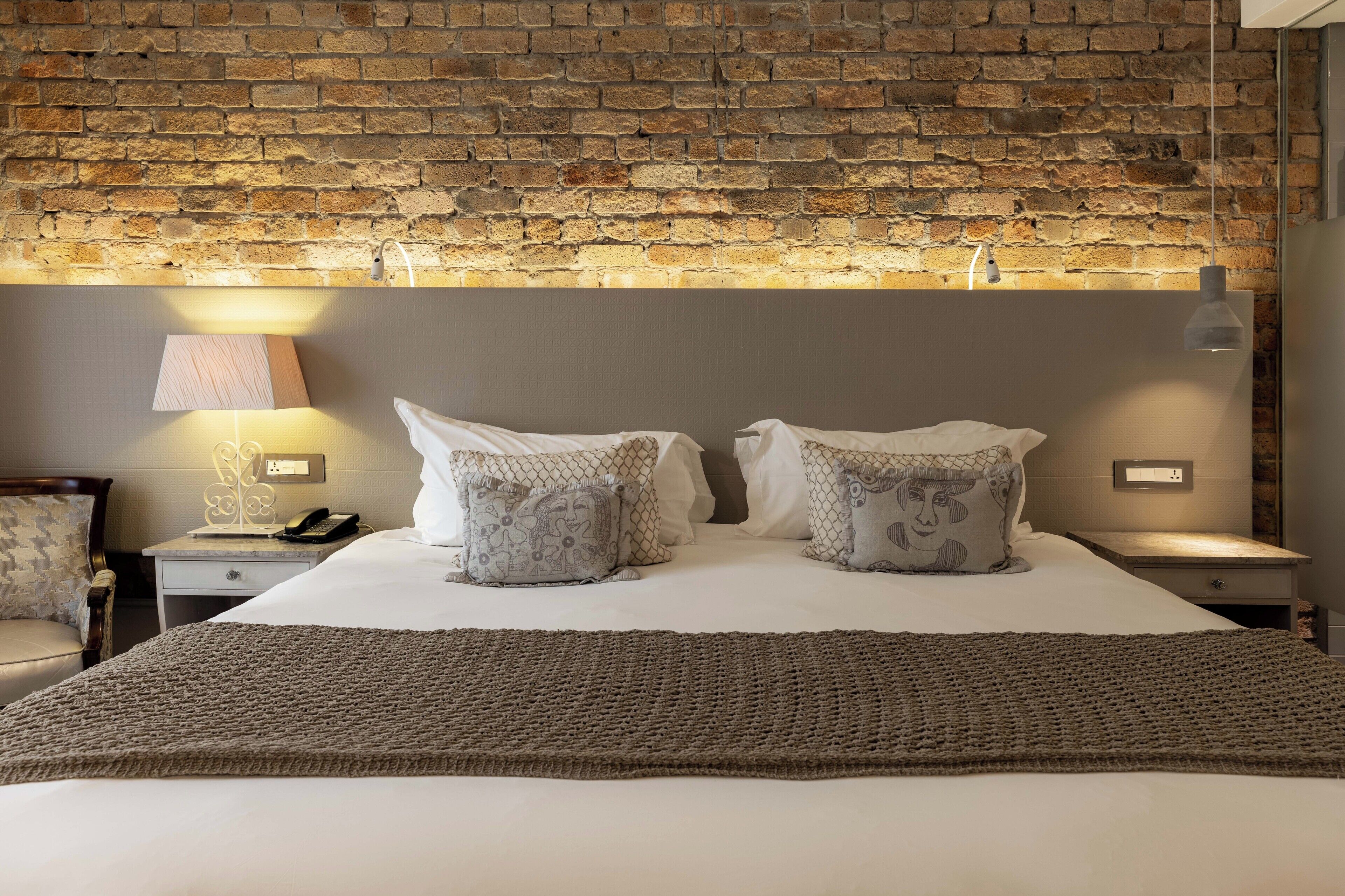 Una habitación de hotel con una pared de ladrillo, una cama con ropa blanca, dos mesitas de noche con lámparas y una silla.