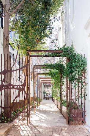 A narrow brick pathway leading to a restaurant with a metal trellis structure and greenery.