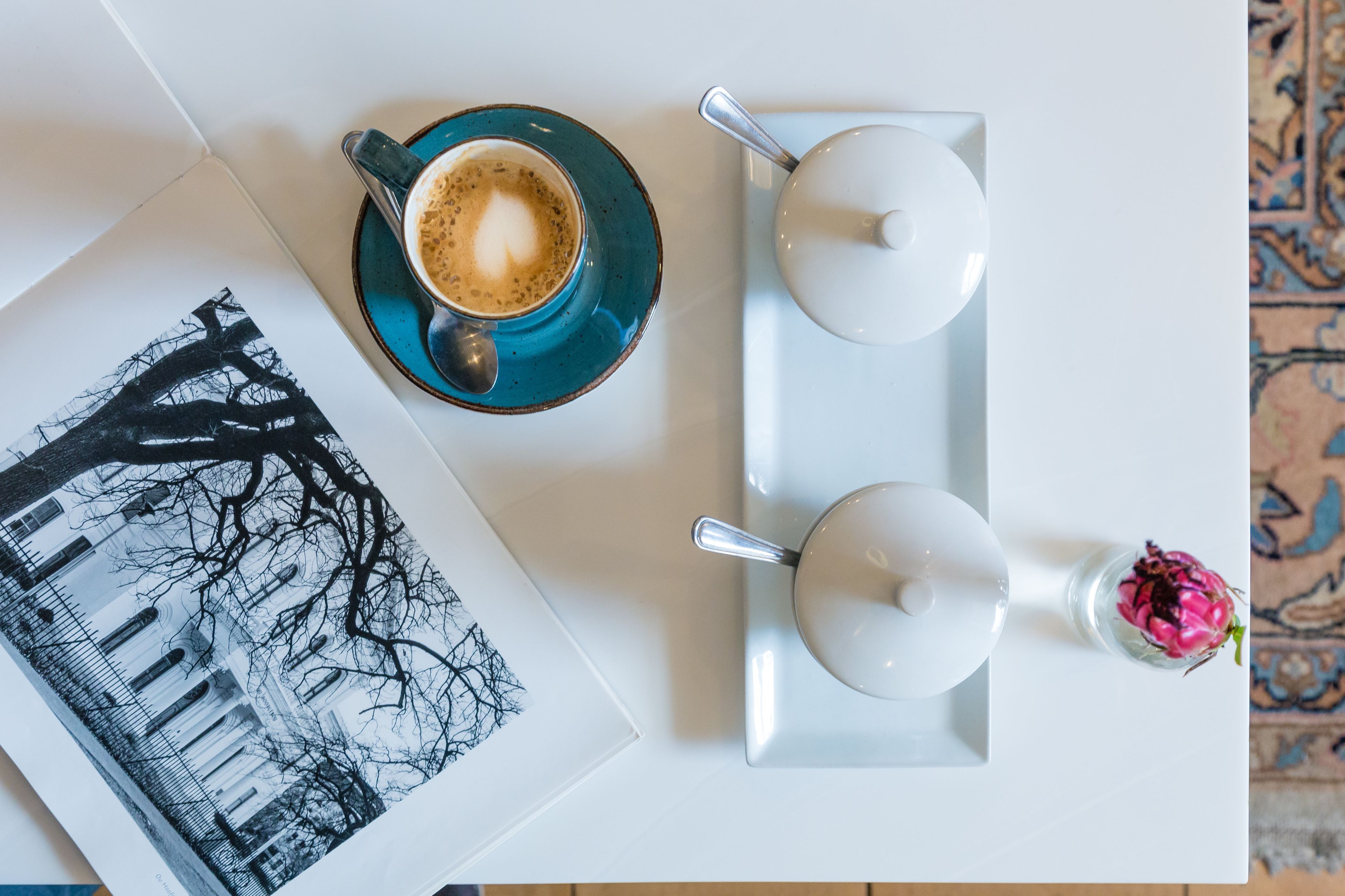 Uma mesa branca com uma xícara de café azul, duas bules brancas, uma colher, um copo de bebida rosa e uma fotografia em preto e branco.