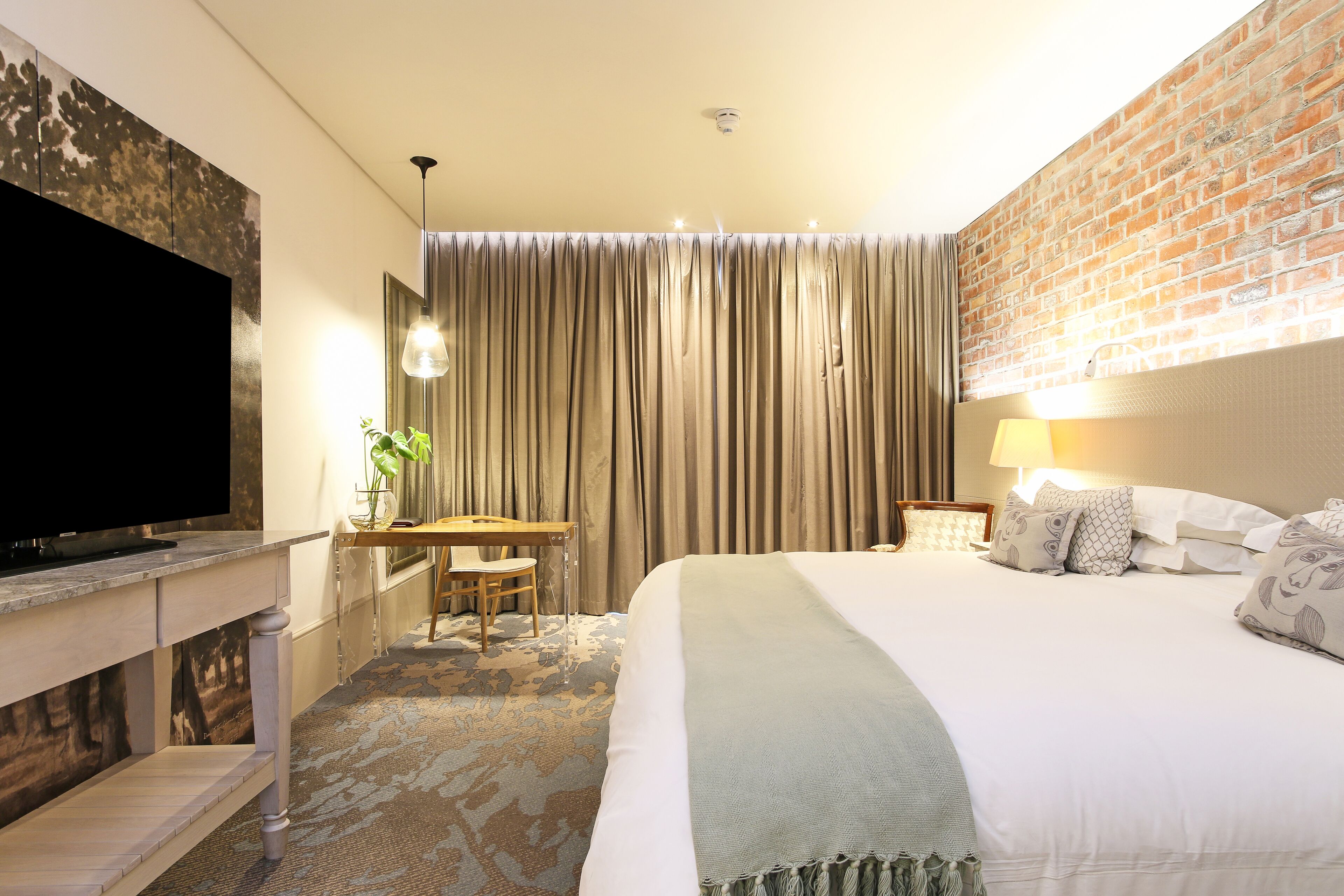 A hotel room with a large flat-screen TV, a desk with a chair, a bed with multiple pillows, and a brick wall.