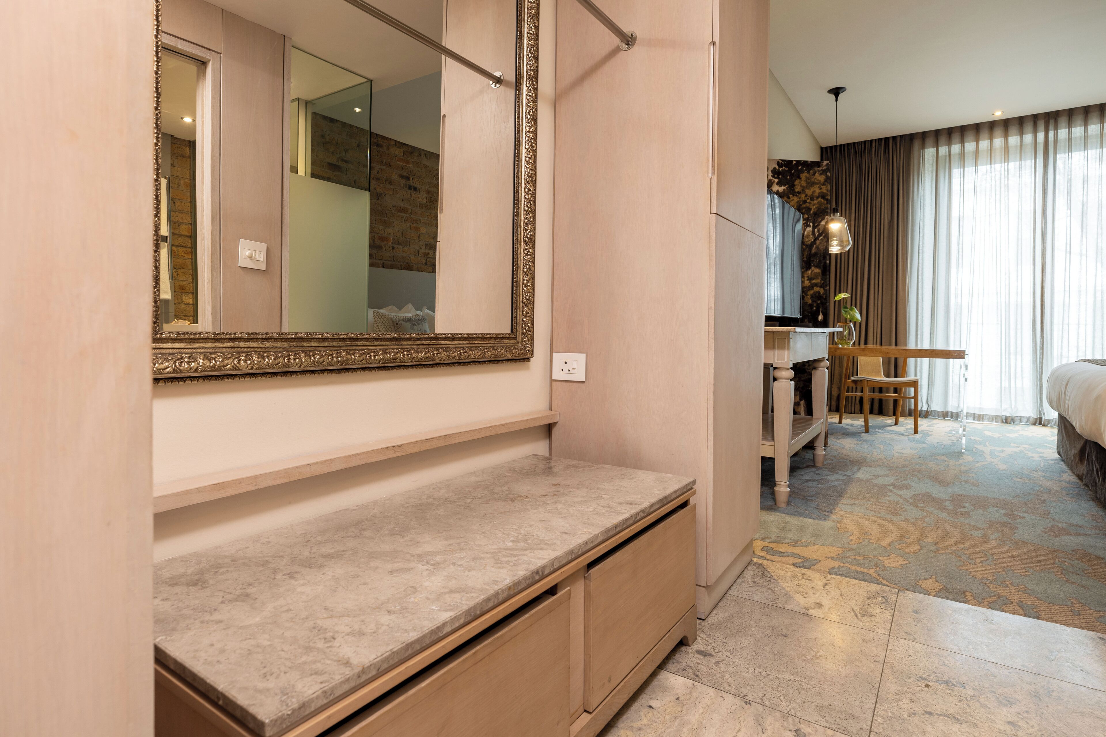 Une chambre d’hôtel moderne dotée d’un grand miroir, d’une coiffeuse avec un lavabo, d’un lit et d’une grande fenêtre avec des rideaux.