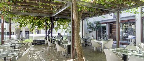Un área de comedor al aire libre con mesas y sillas, una pérgola con enredaderas y un árbol en el centro.