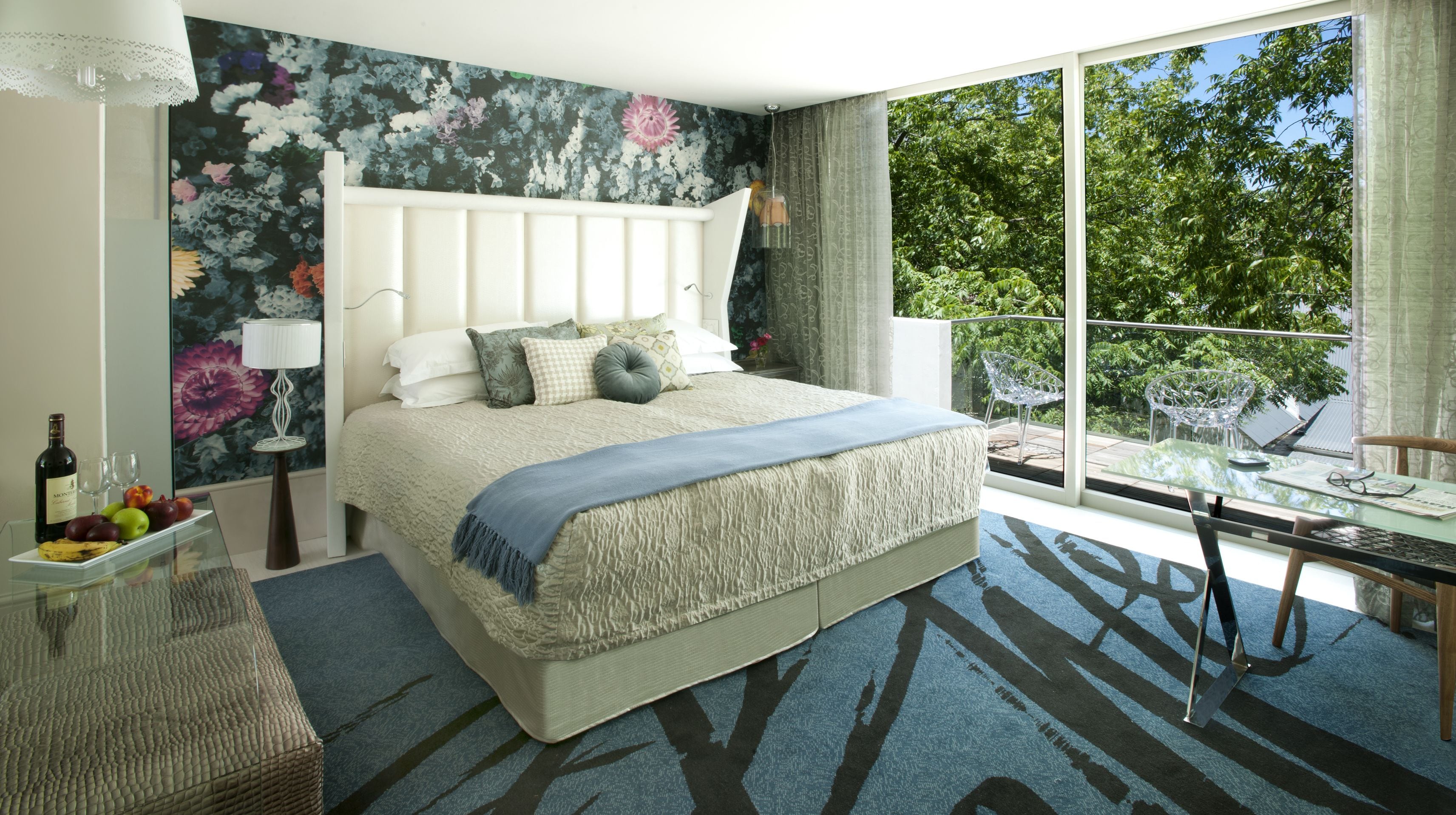 Un dormitorio con una cama grande, una mesa de vidrio con una silla, una mesita de noche con lámpara y vistas a árboles por la ventana.