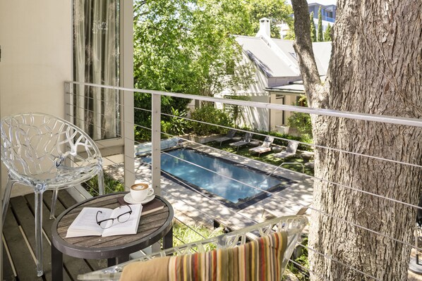 Un balcón con vista a una piscina, una silla, una mesa con un libro y vasos, y un árbol.
