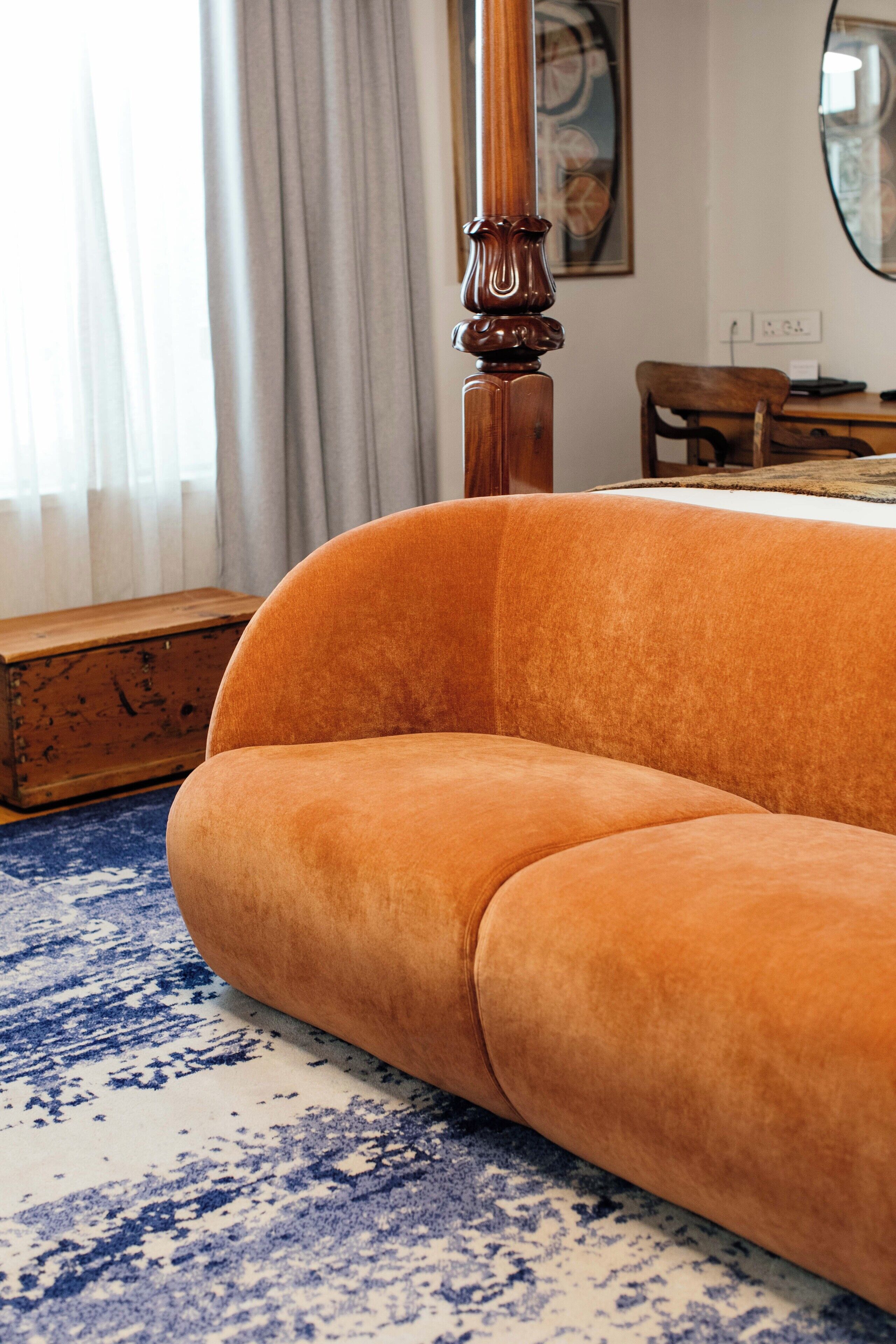 A room with a large orange sofa, a wooden headboard, a wooden chest, and a patterned rug.