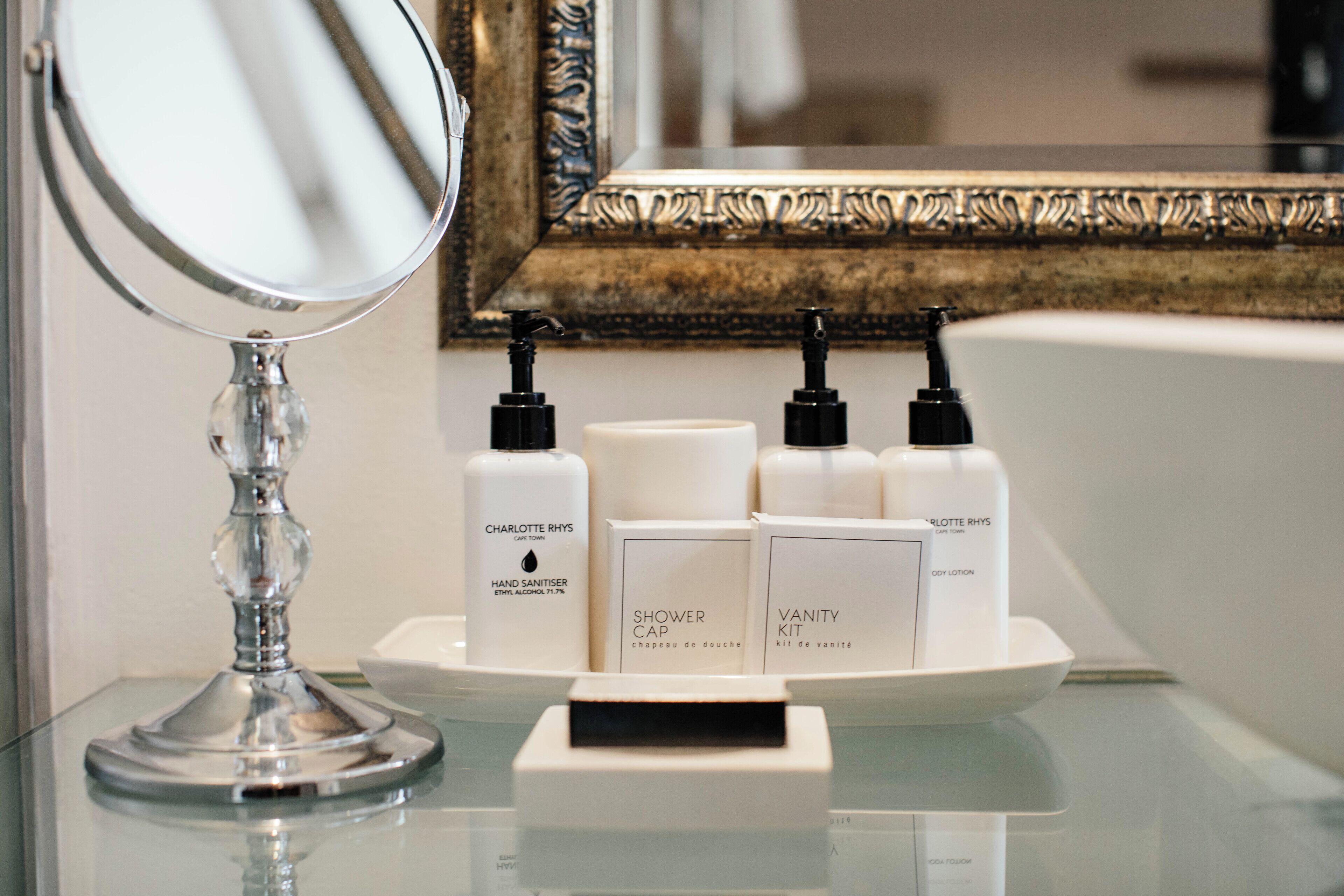 A bathroom vanity with a mirror, a soap dispenser, a bottle of hand sanitizer, a shower cap, and a vanity kit.