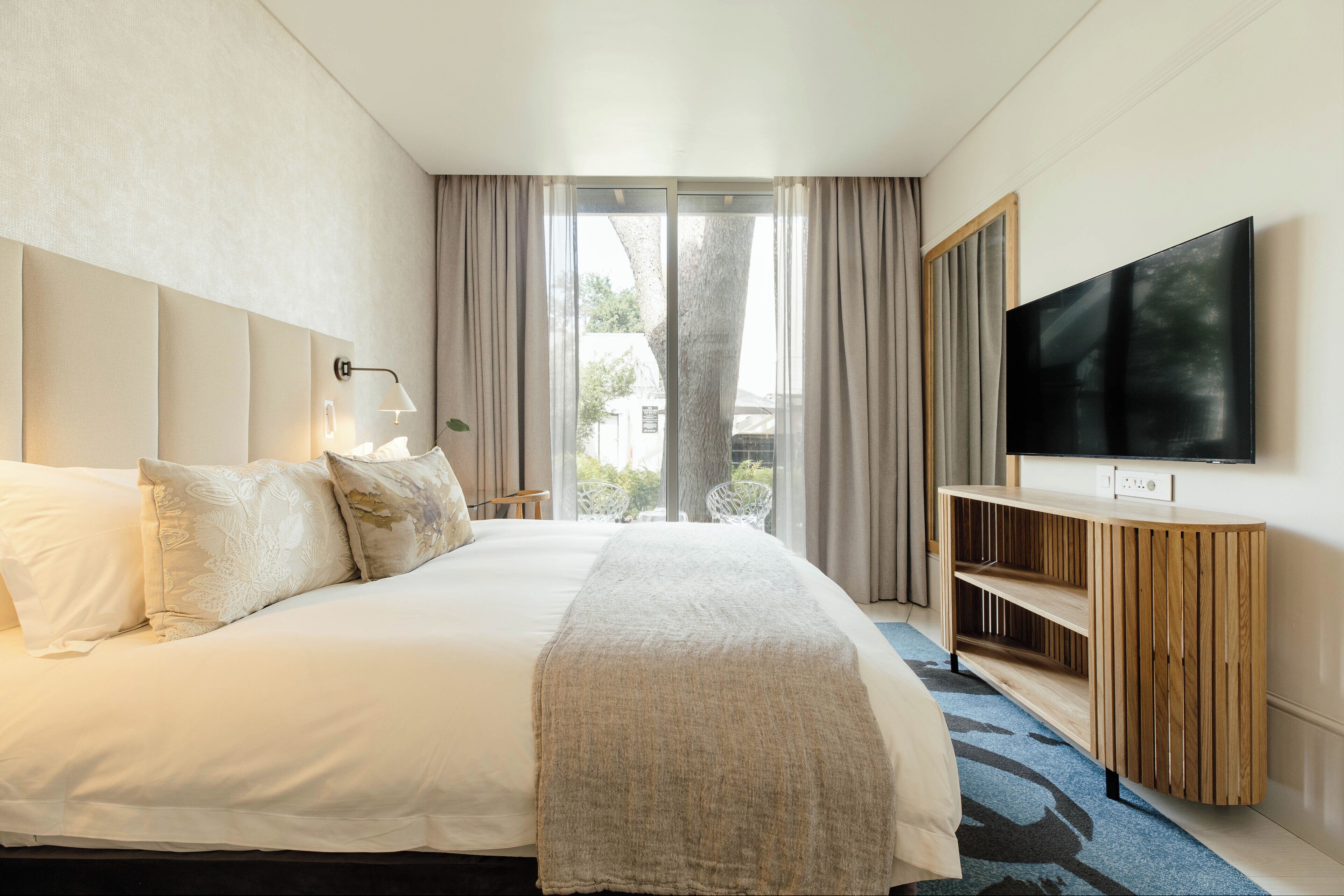 A hotel room with a large bed, a TV on a wooden console, and a view of the outdoors through a sliding door.