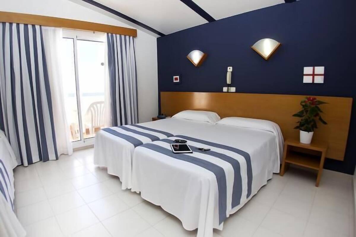 Ein Hotelzimmer mit zwei Betten, einem großen Fenster mit Vorhängen, einer Wandleuchte und einer kleinen Pflanze auf einem Nachttisch.
