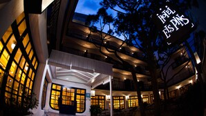 Entrada de hotel con área de estar al aire libre techada, varios balcones y un letrero iluminado destacando.