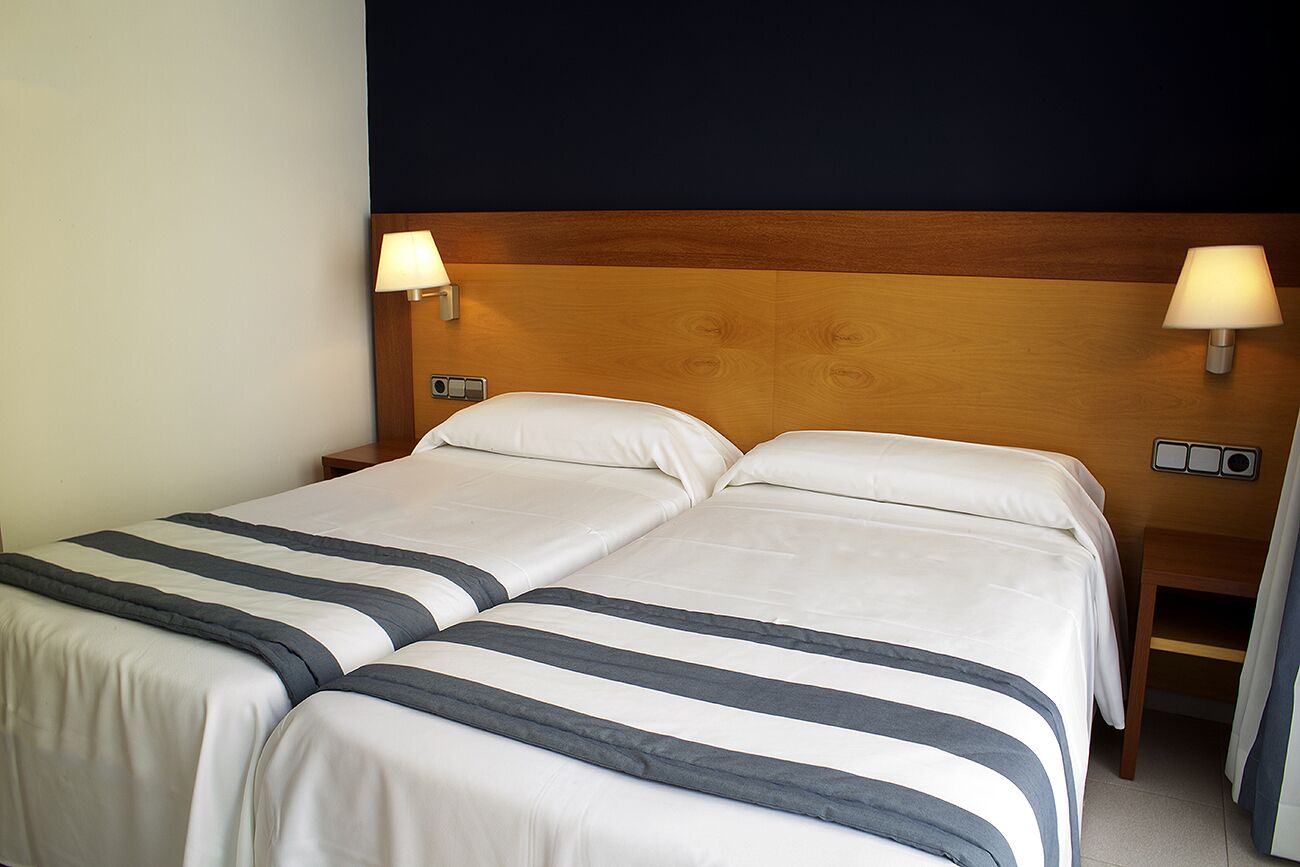 Habitació d'hotel amb dos llits, cadascun amb capçalera de fusta, roba de llit blanca i abernissadors.
