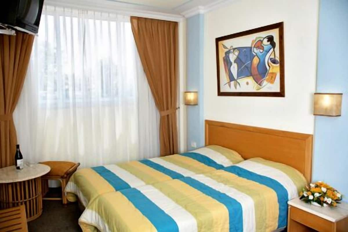 A hotel room with a bed, a small table with a bottle and glasses, a chair, a painting on the wall, and a nightstand with a vase of flowers.