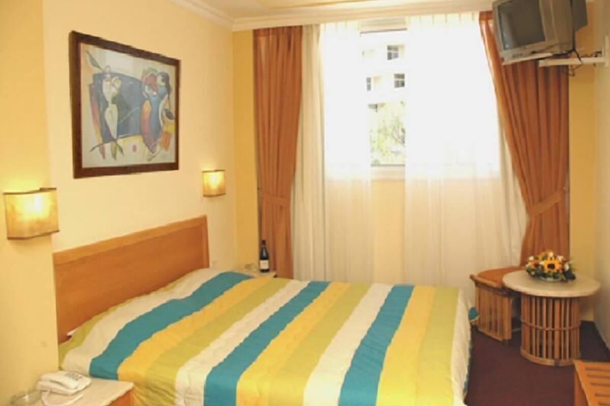 A hotel room with a bed, a television mounted on the wall, a small round table with a flower arrangement, and a painting on the wall.