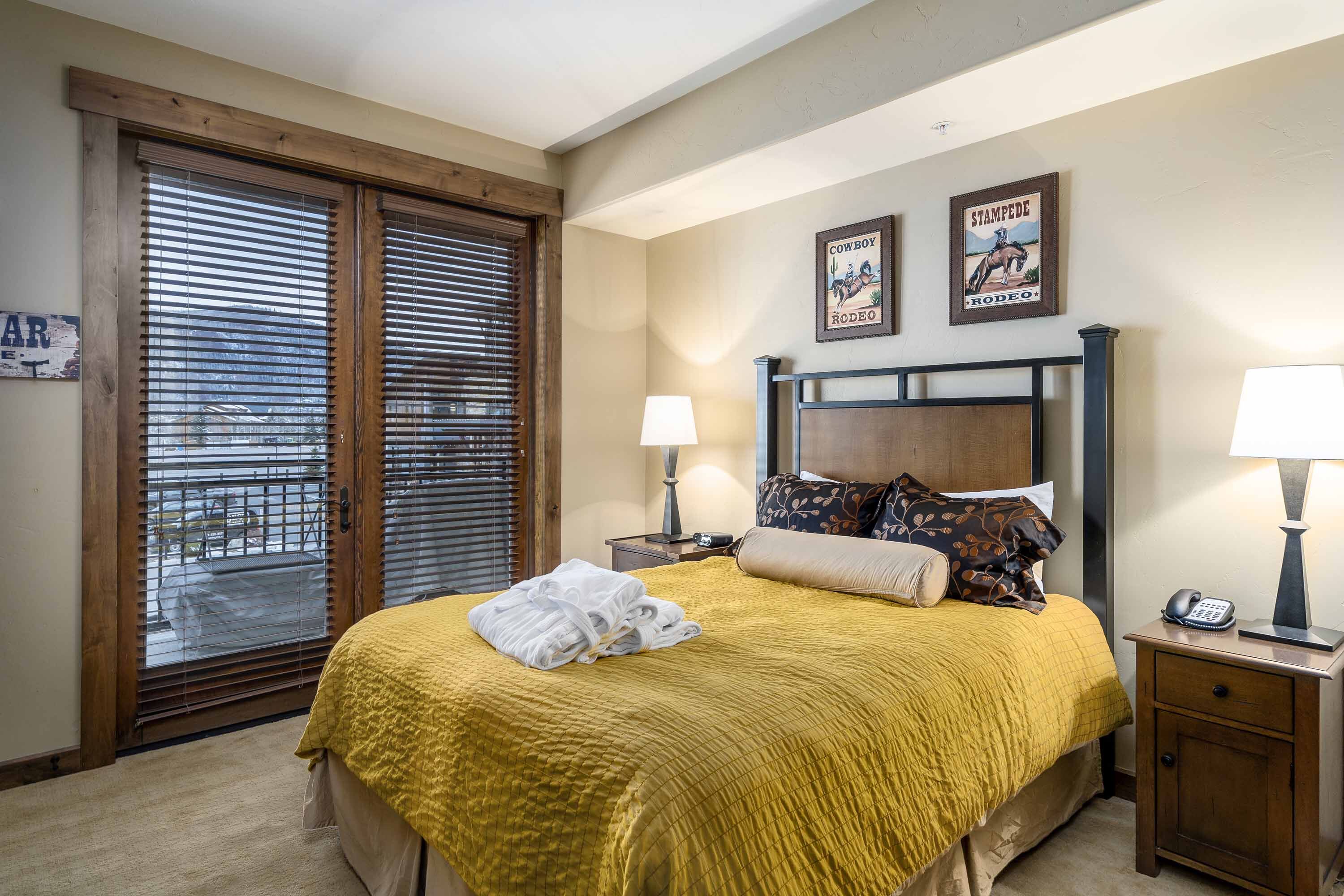 A hotel room with a bed, two bedside lamps, a nightstand, a door with blinds, and two wall decorations.