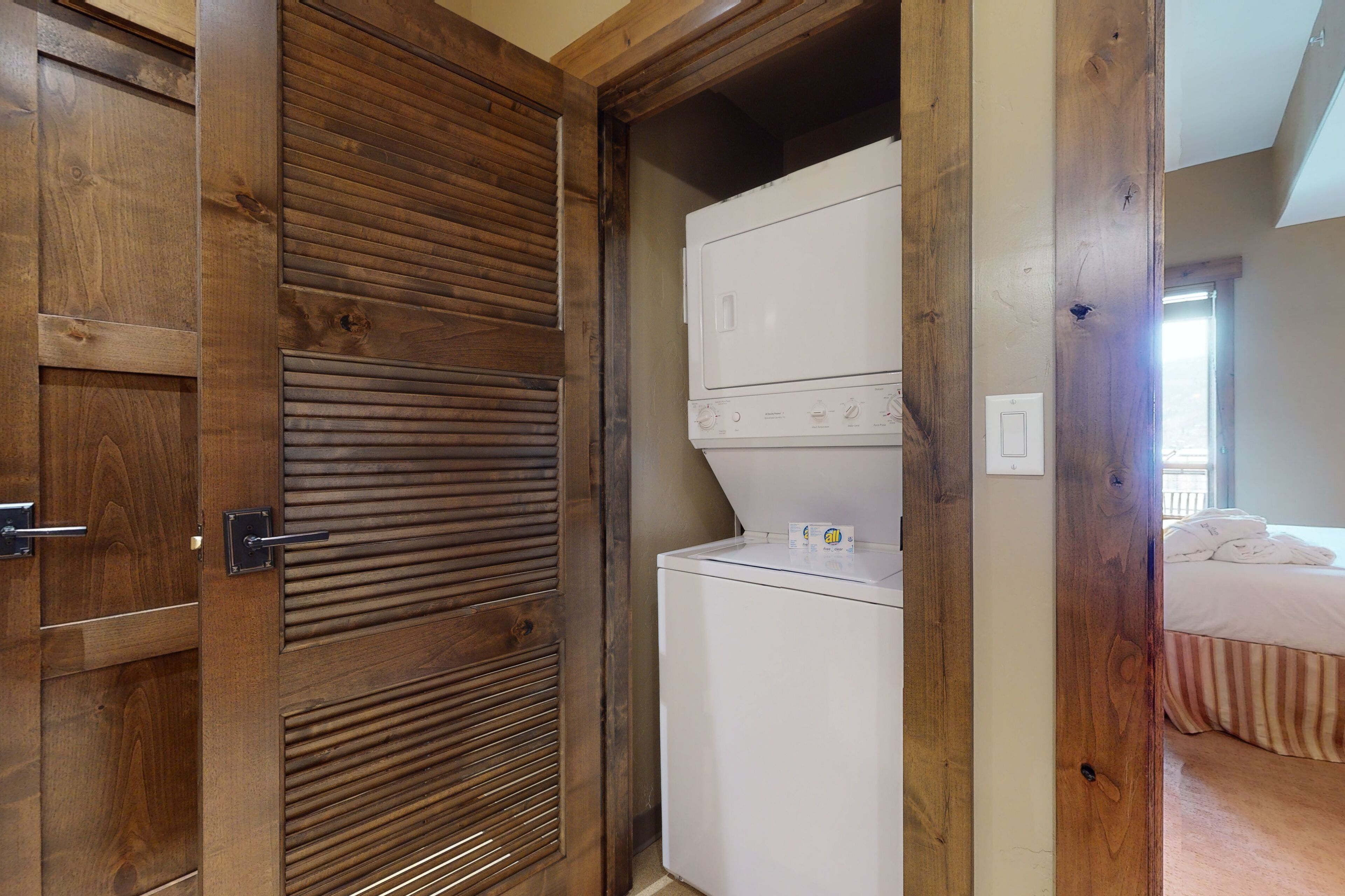 A laundry room with a white washer and dryer, wooden doors, and a bed in the adjacent room.