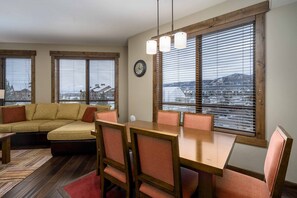 A cozy living room with a sectional sofa, a dining table with chairs, large windows with blinds, and a view of a snowy landscape.
