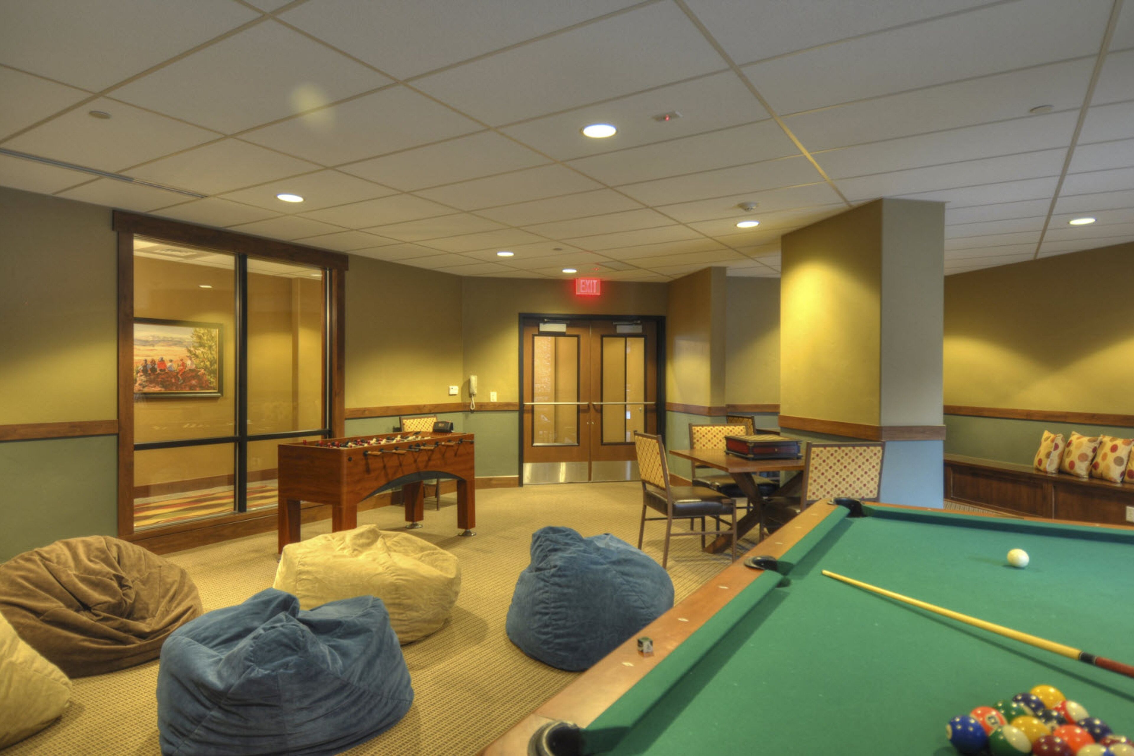 A pool table with billiard balls, bean bags, and a foosball table in a room with a door and a wall-mounted picture.