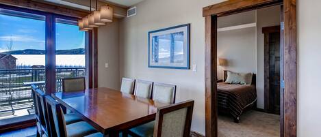 A dining area with a wooden table and chairs, a large window with a view, and a framed picture on the wall.