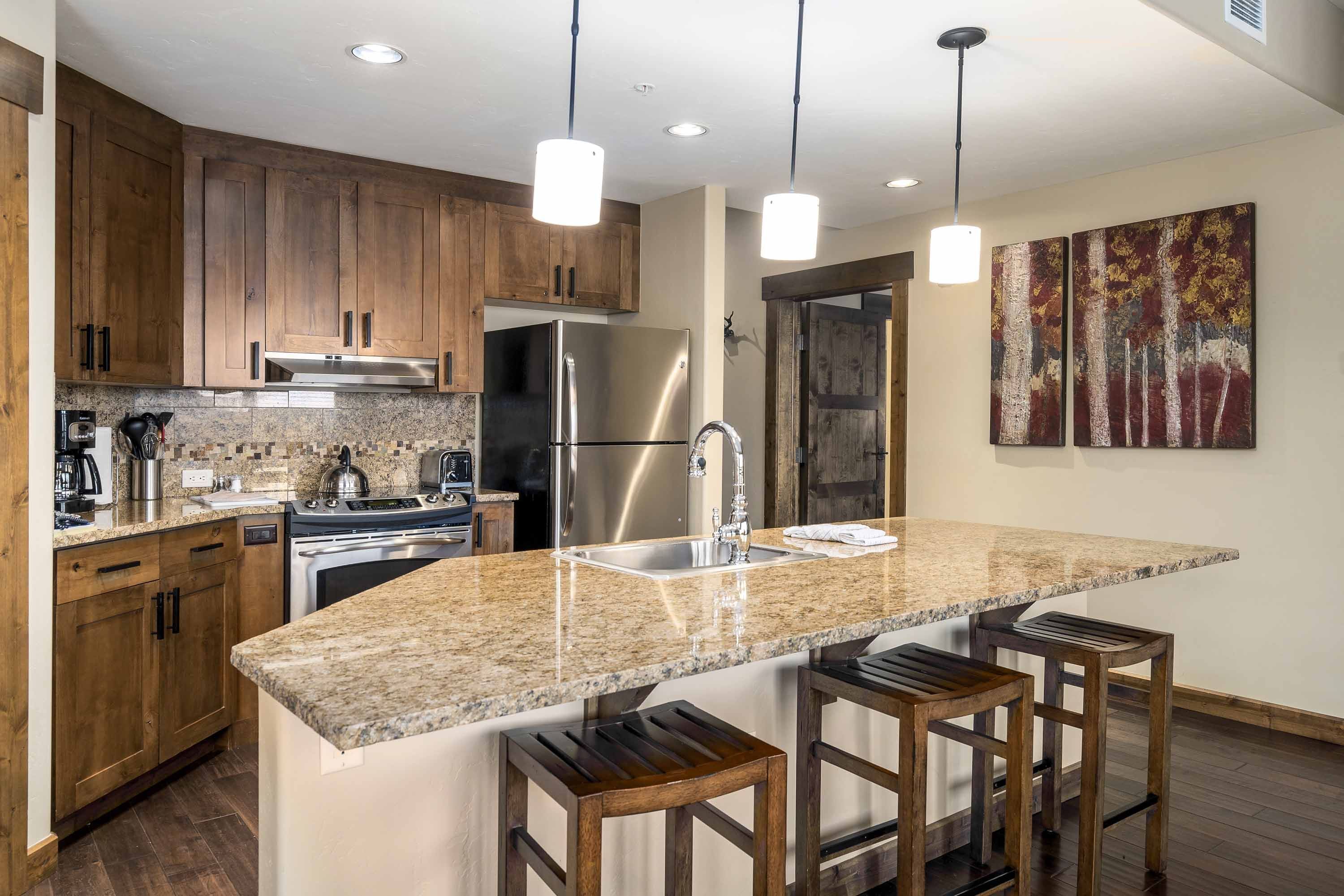 Una cocina moderna con encimeras de granito, electrodomésticos de acero inoxidable, armarios de madera e iluminación colgante.