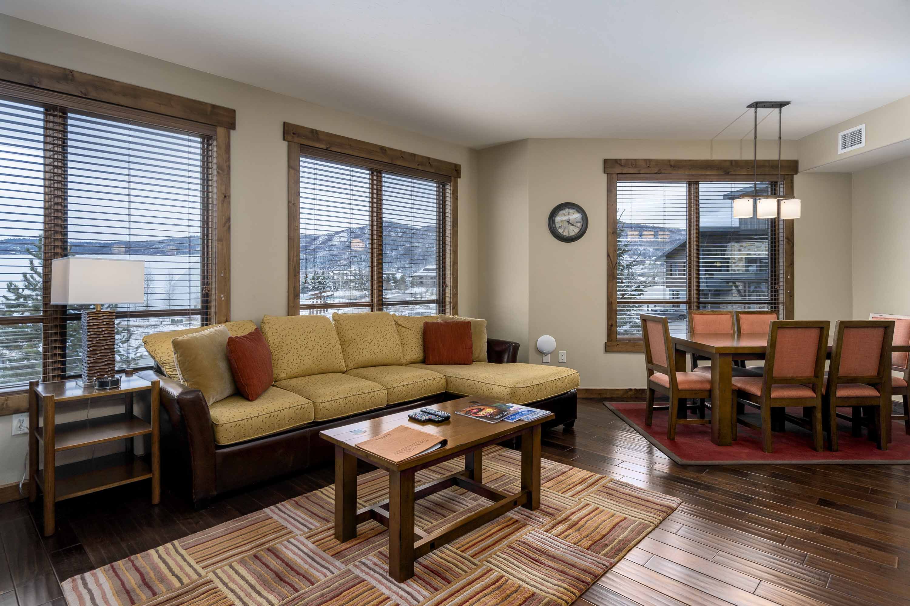 A living room with a sofa, coffee table, dining table with chairs, and large windows with blinds.