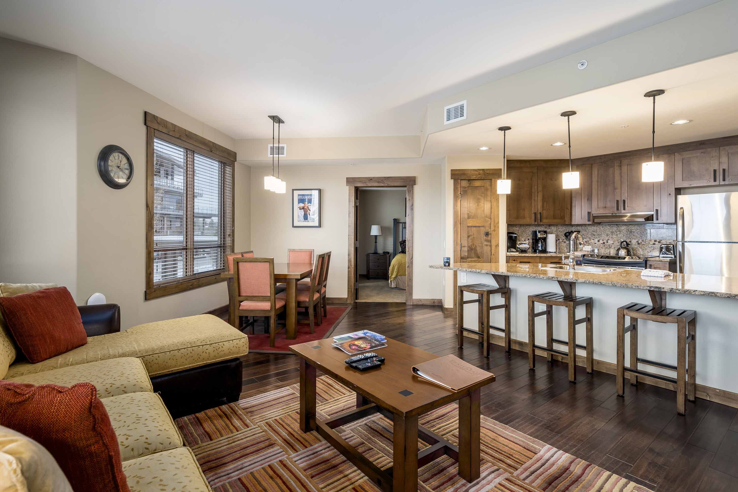 A modern living room with a sofa, coffee table, and dining area. It features a kitchen with wooden cabinets and bar stools.