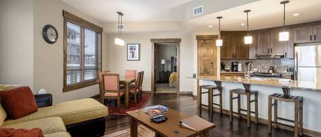 A modern living room with a sofa, coffee table, and dining area. It features a kitchen with wooden cabinets and bar stools.