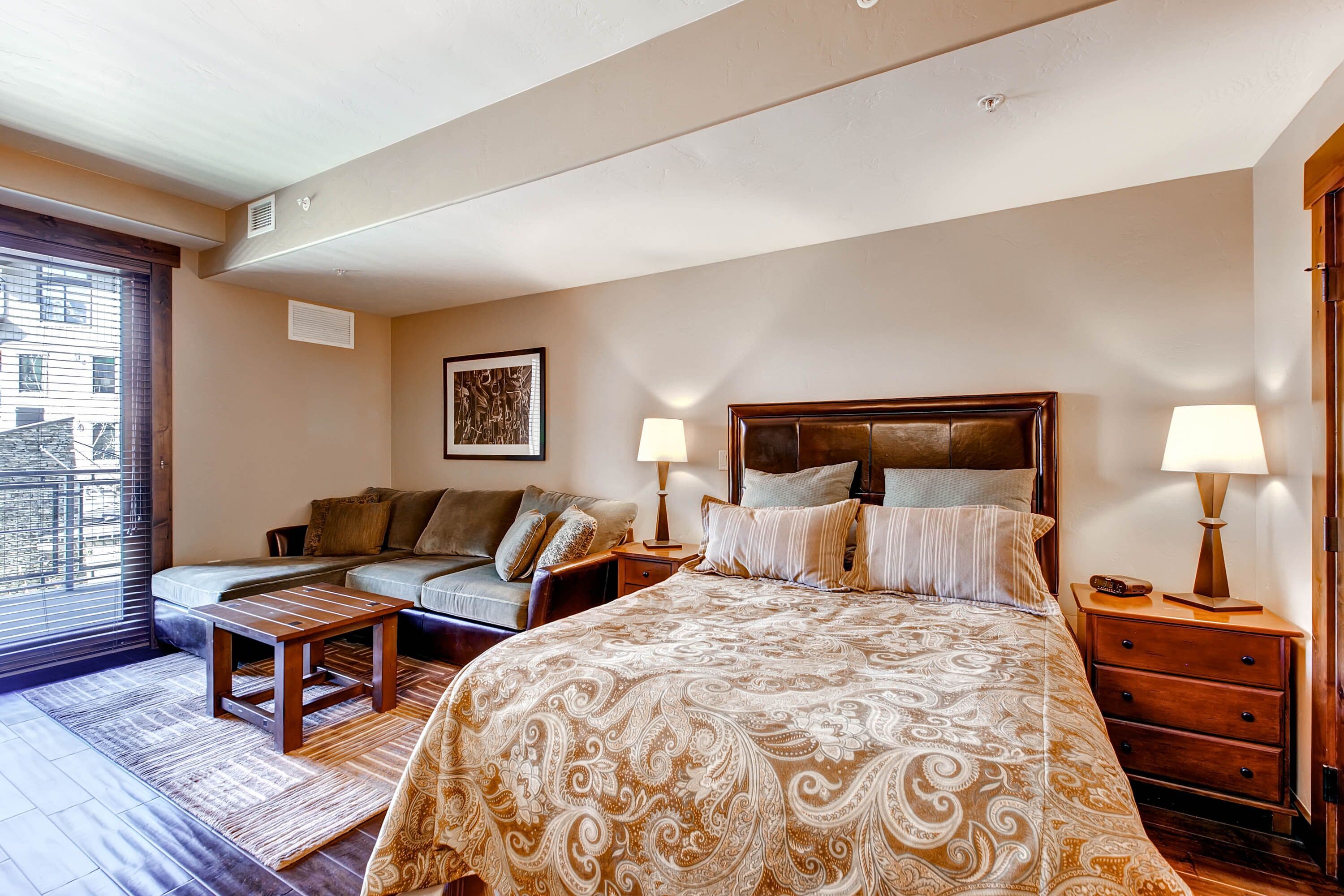 A bedroom with a large bed, a sofa, a small table, two bedside tables with lamps, and a window with blinds.