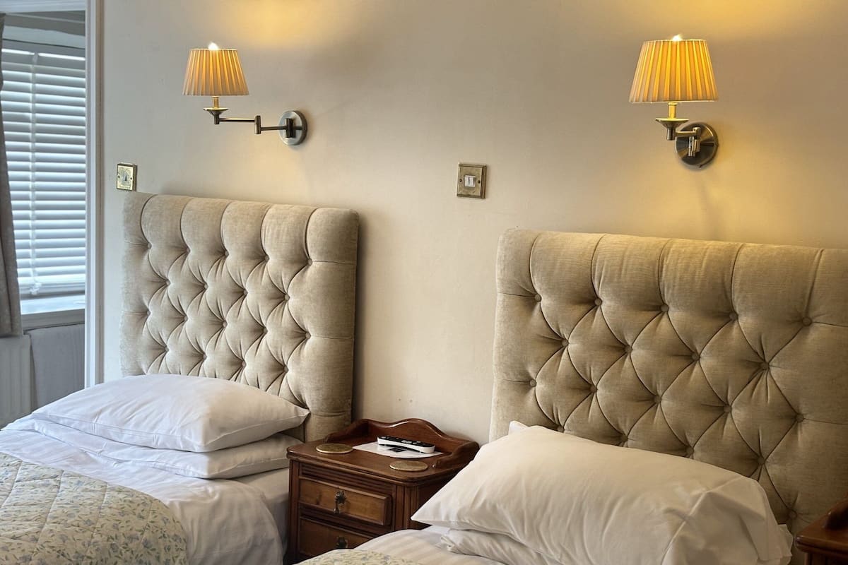 A hotel room with two beds, each with a tufted headboard, bedside tables, lamps, and a window with blinds.