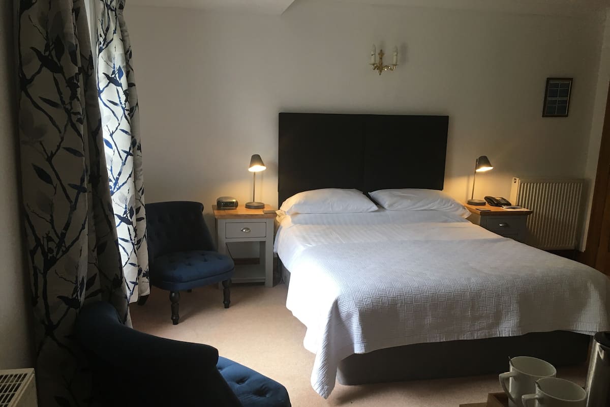 A hotel room with a bed, two bedside tables, a chair, a desk with a lamp, and a radiator.