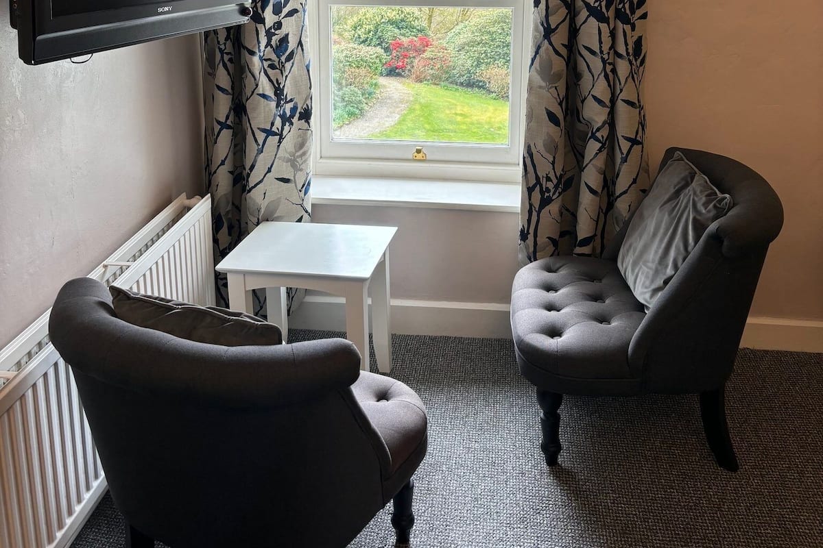 A room with a flat-screen TV mounted on the wall, a small white table, two dark armchairs, and a window with patterned curtains.