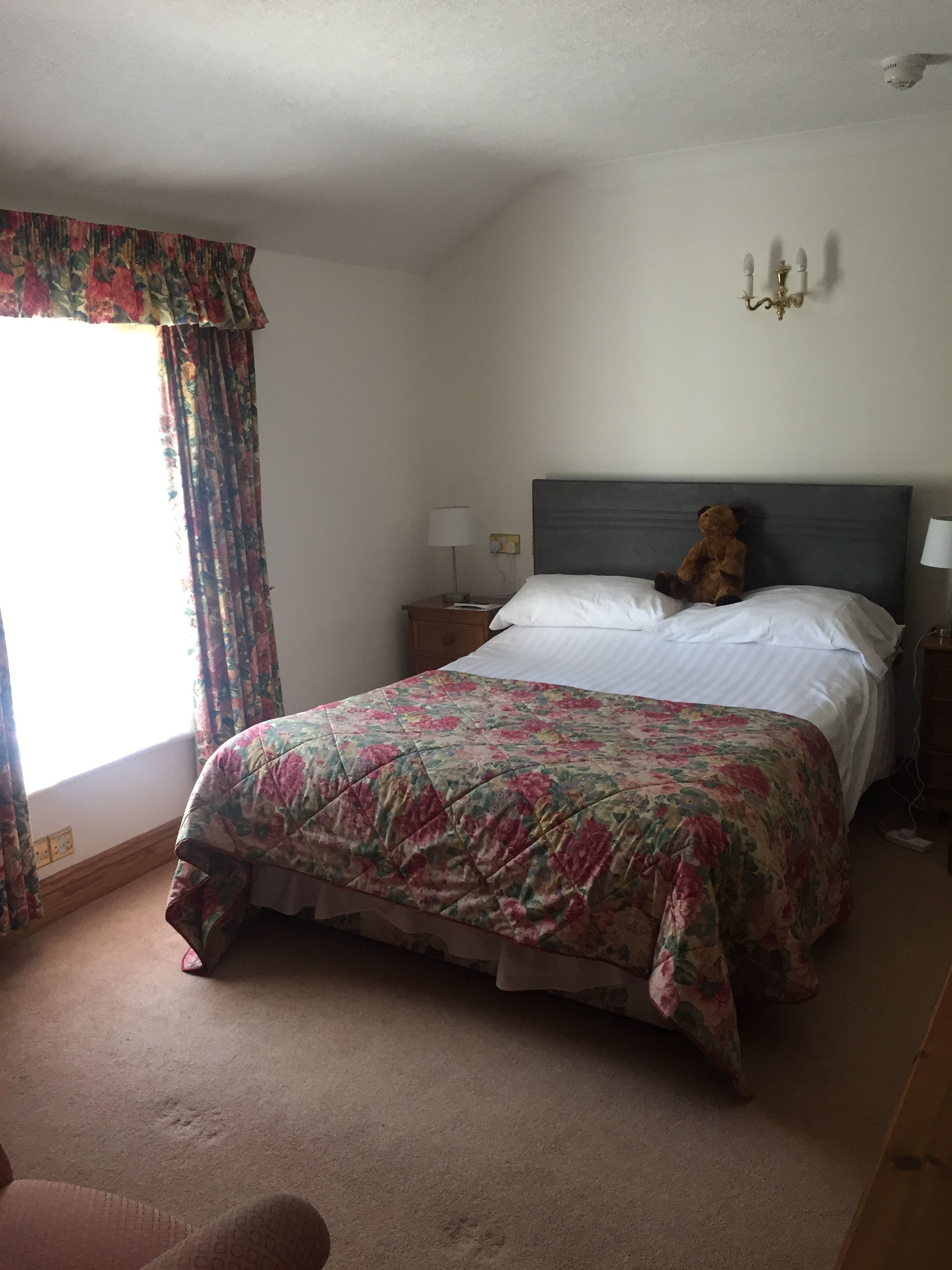 A neatly made bed with a floral bedspread, a bedside lamp, and a teddy bear on the headboard.