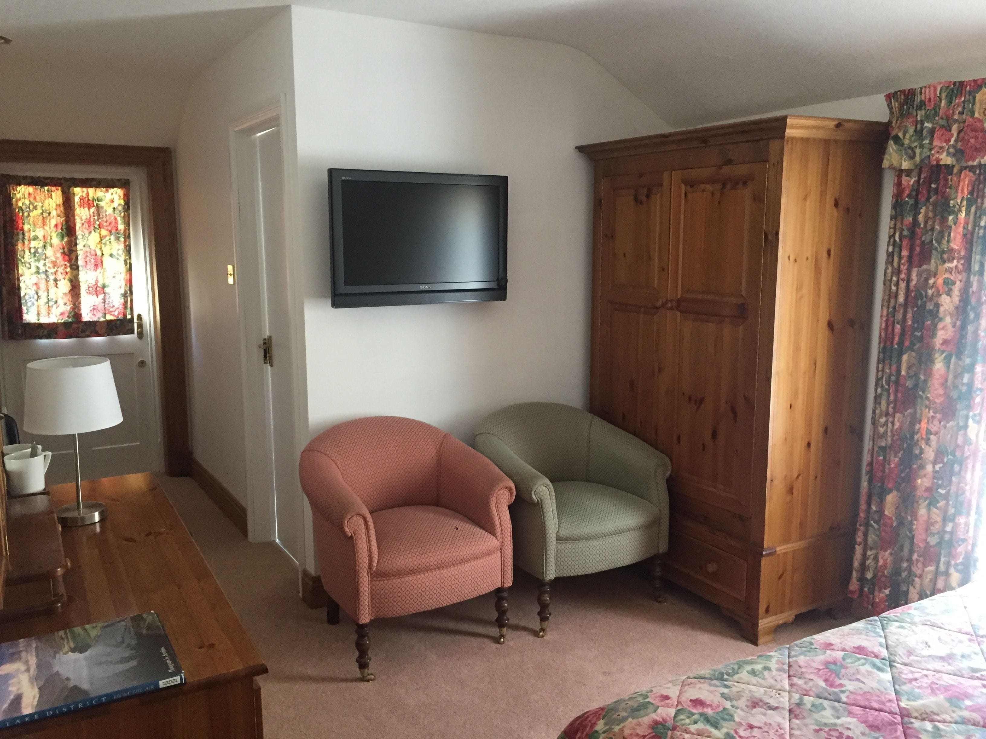 A room with a wooden wardrobe, two chairs, a TV mounted on the wall, and a door with floral curtains.