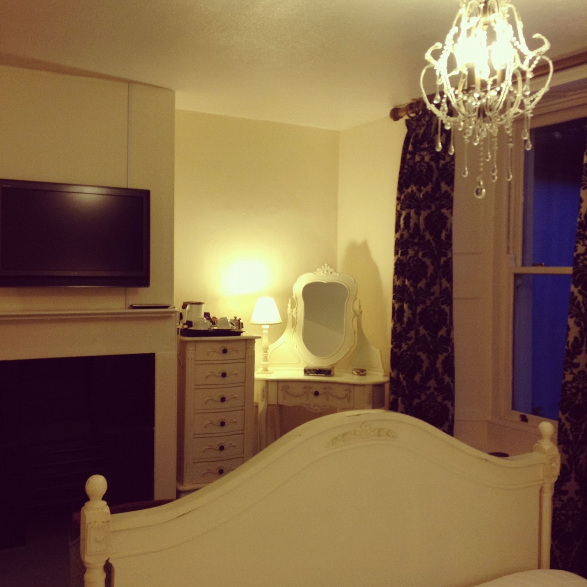 A bedroom with a bed, dresser, vanity with mirror, chandelier, and a television on the wall.