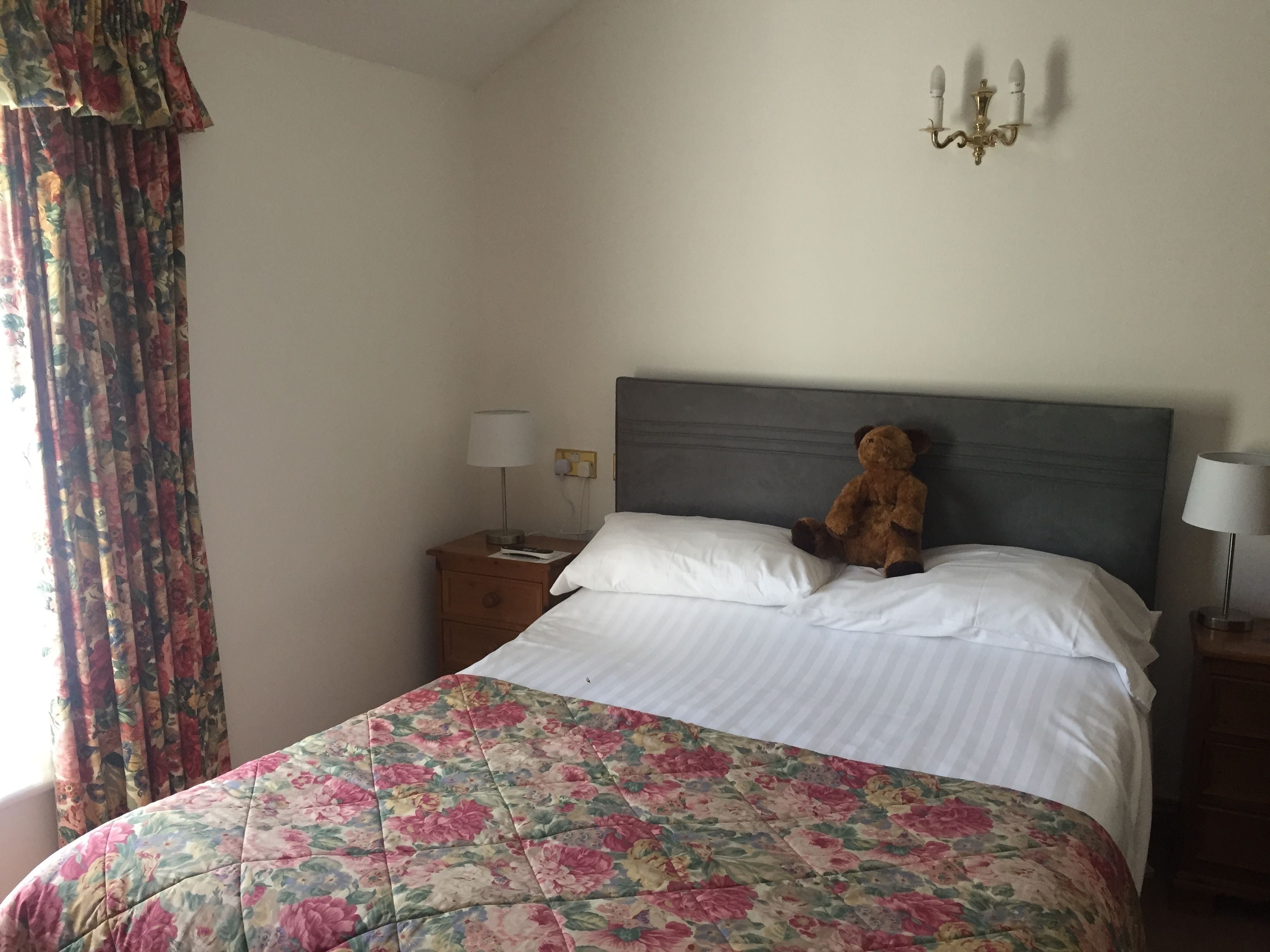 A neatly made bed with a floral bedspread, a bedside lamp, a nightstand, and a teddy bear on the bed.