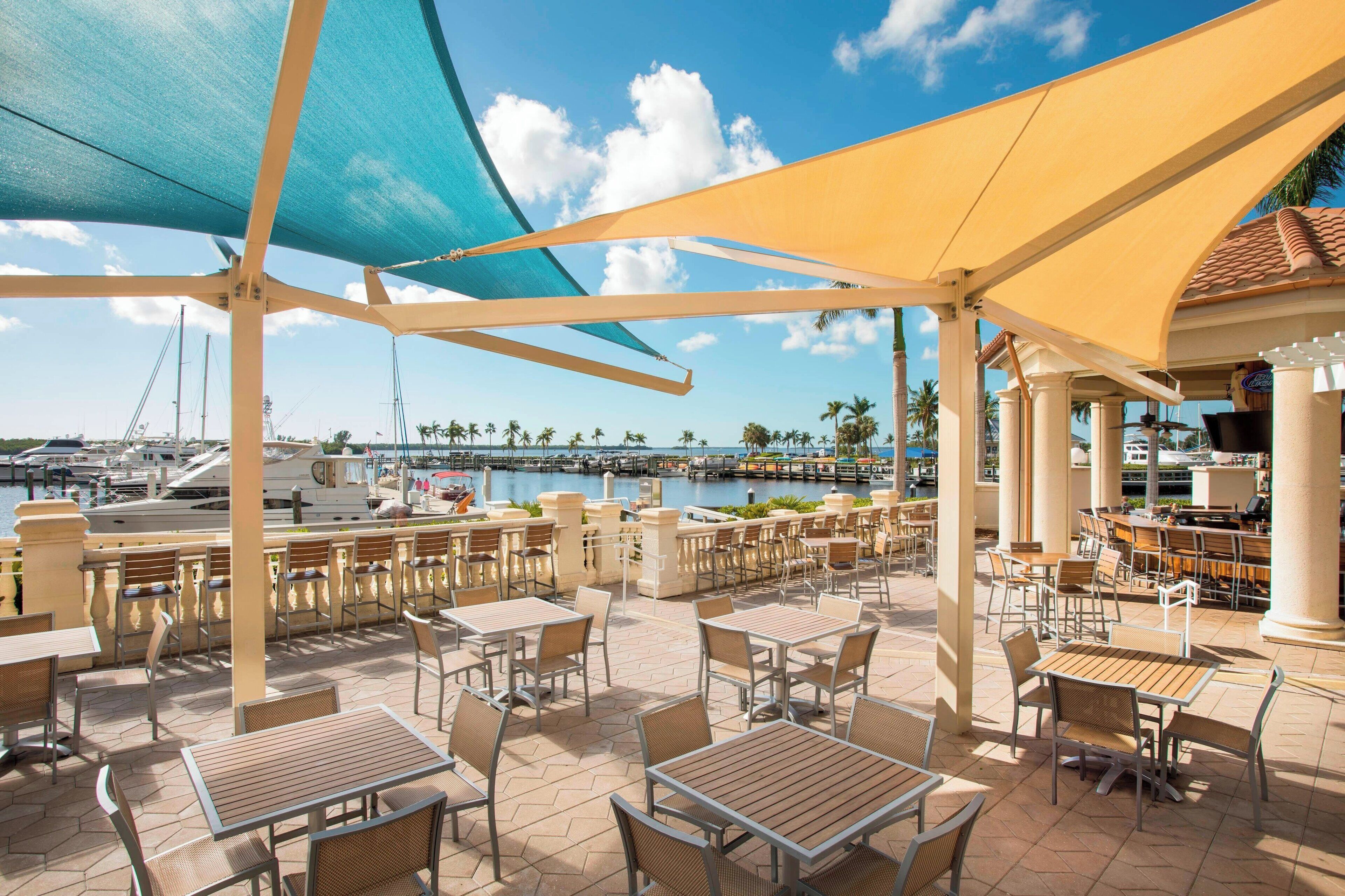 An outdoor patio with tables and chairs, overlooking a marina with boats and palm trees.