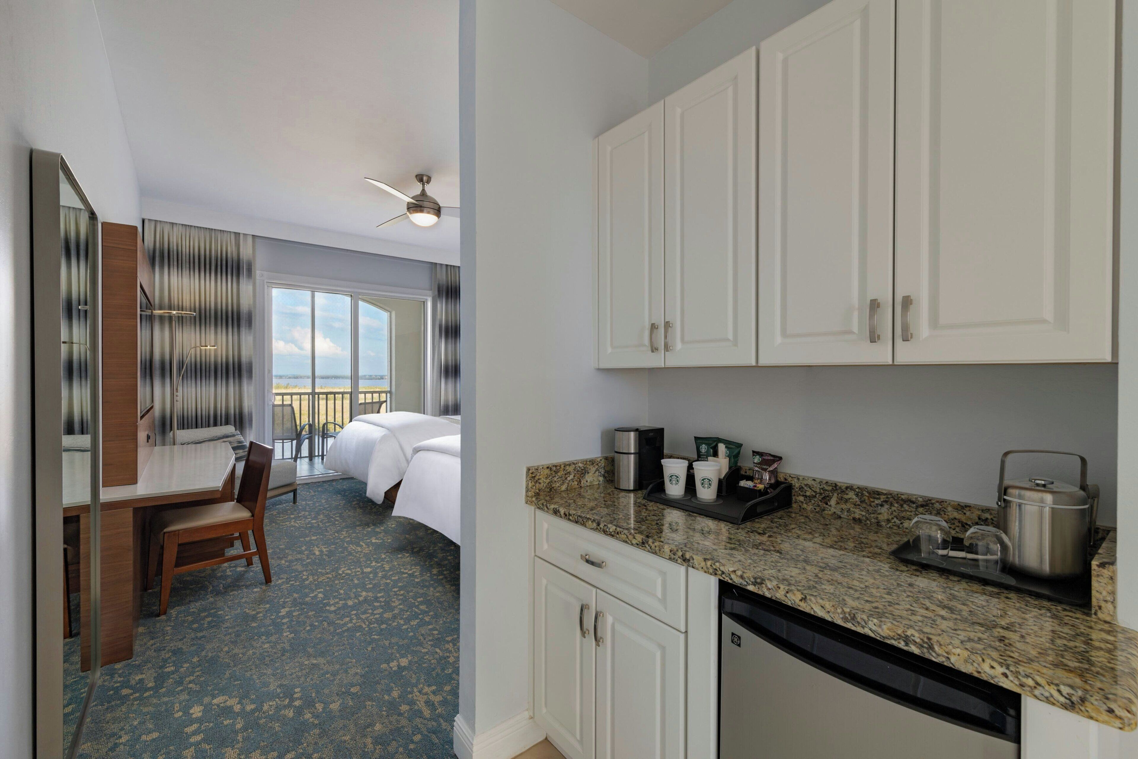 A hotel room with a bed, a desk, a chair, a kitchenette with a coffee maker, and a view of the sky.