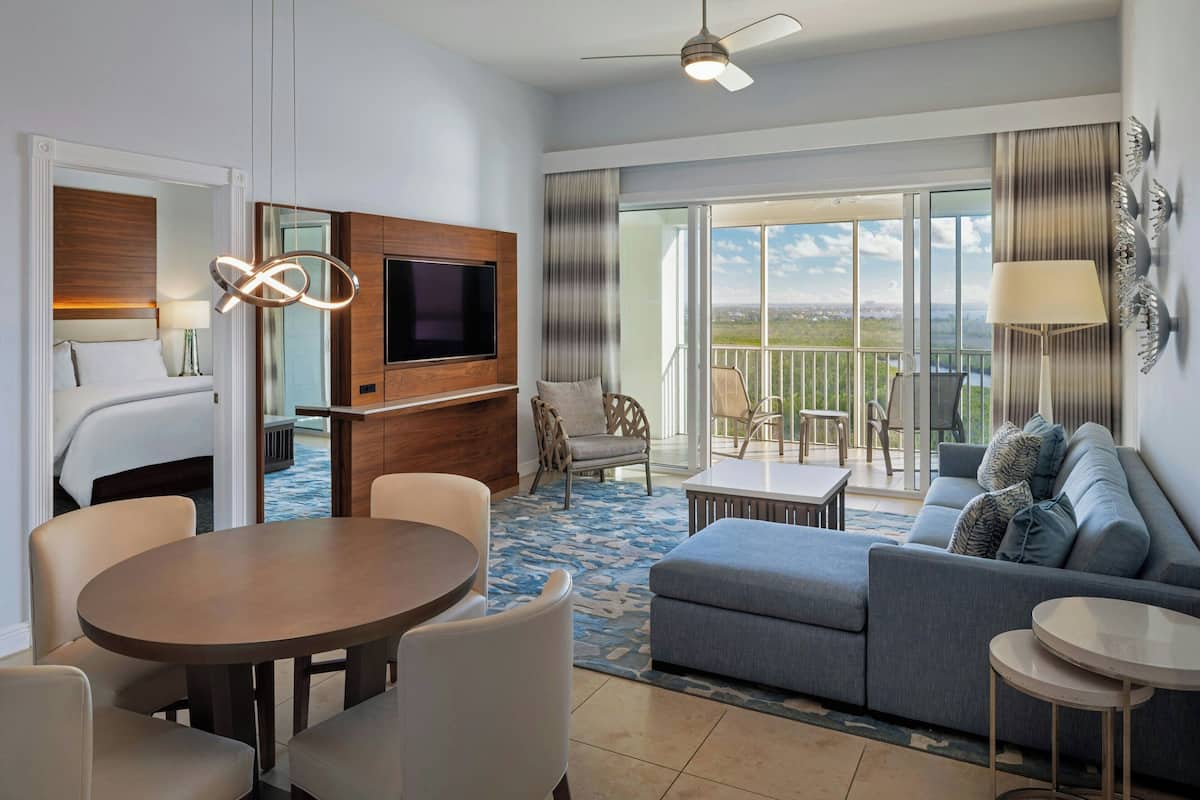 Quarto de hotel com varanda, área de refeições, sofá e televisão.