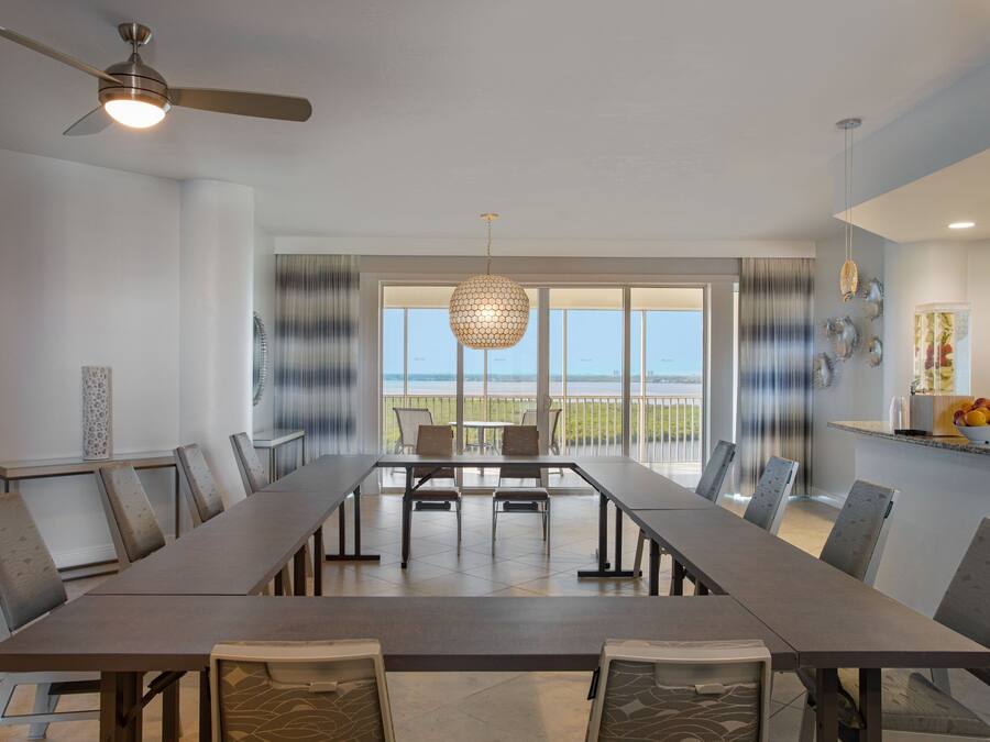 A spacious dining area with a long table and chairs, a ceiling fan, and a view through large windows to the outdoors.