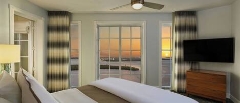 Habitación de hotel con cama, televisor, ventilador de techo y vista al atardecer por la ventana.