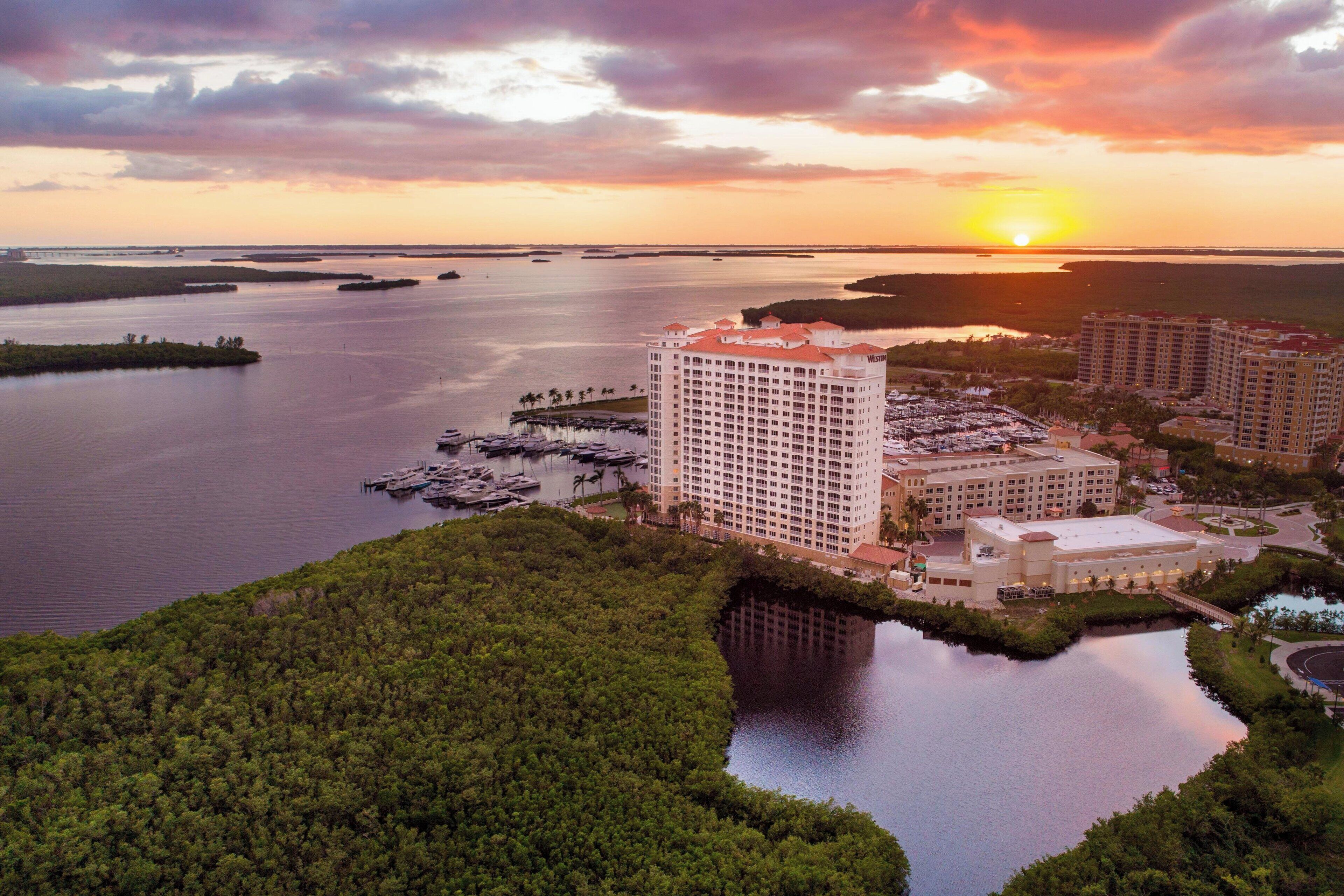 A hotel with multiple buildings, a marina with boats, and a sunset over a body of water.